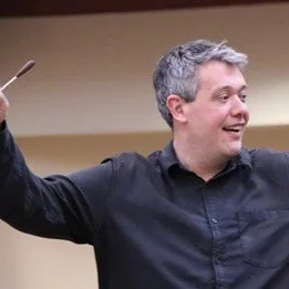 A man conducting an orchestra or choir, holding a baton in his right hand, smiling and looking to his left.