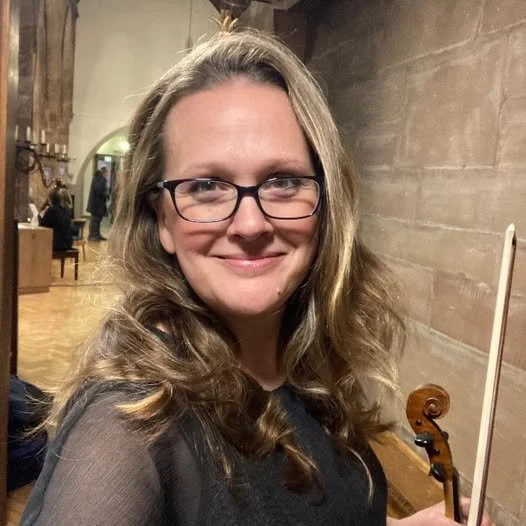 A woman wearing glasses and a black top, holding a violin, smiling, inside a room with wooden walls and a rustic interior.