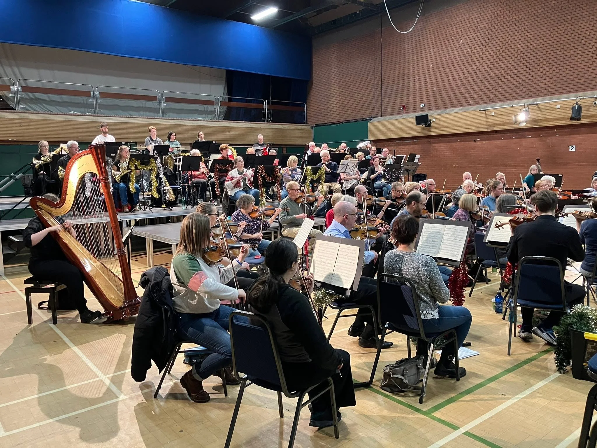 Orchestra performing in a gymnasium decorated with holiday garlands and ornaments, with musicians playing violins, flutes, and other instruments.