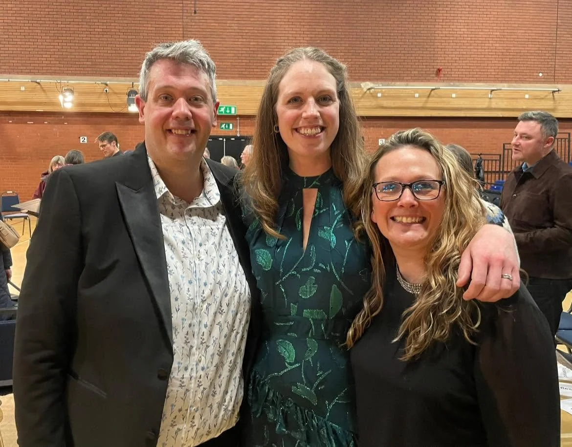 Three people smiling and posing together at an indoor event, with a brick wall and other attendees in the background.