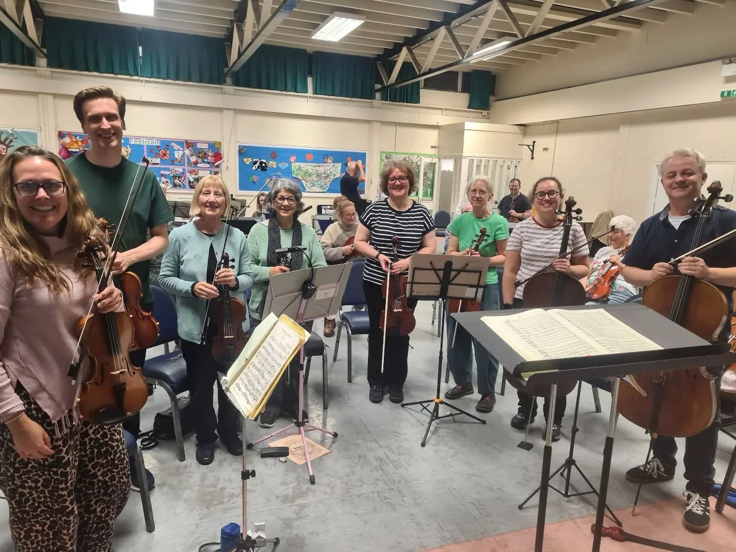 A group of ten musicians holding string instruments, including violins and cellos, standing in a rehearsal room with music stands and sheet music, smiling at the camera.