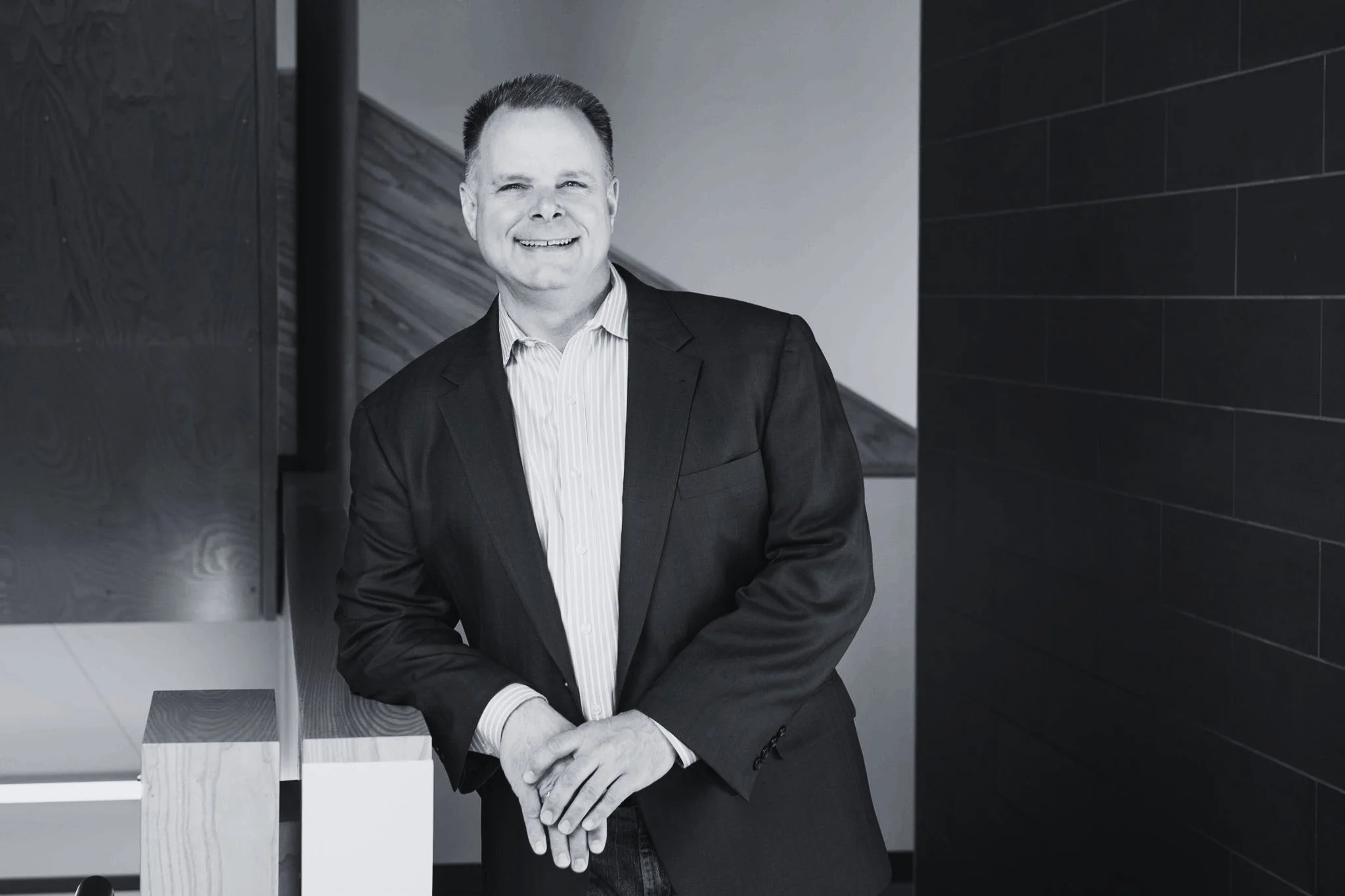 Black and white photograph of a smiling man in a suit standing with his arms crossed in a modern indoor setting.