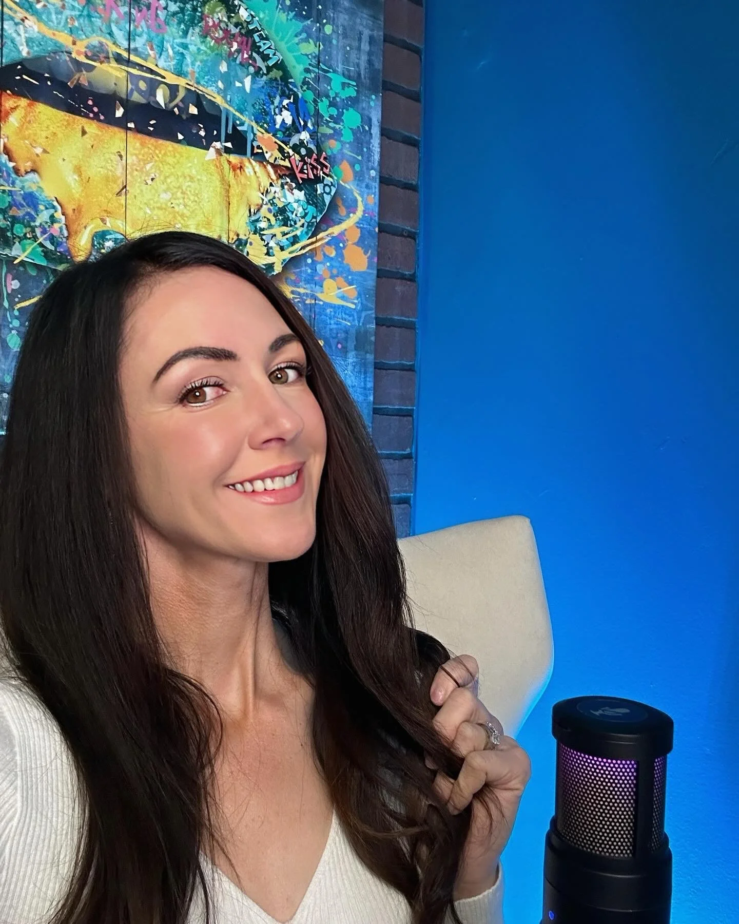 A woman with long dark hair smiling and sitting in front of a colorful abstract art piece. She is wearing a white top and has a silver ring on her finger. There is a black and purple microphone in front of her on the table.