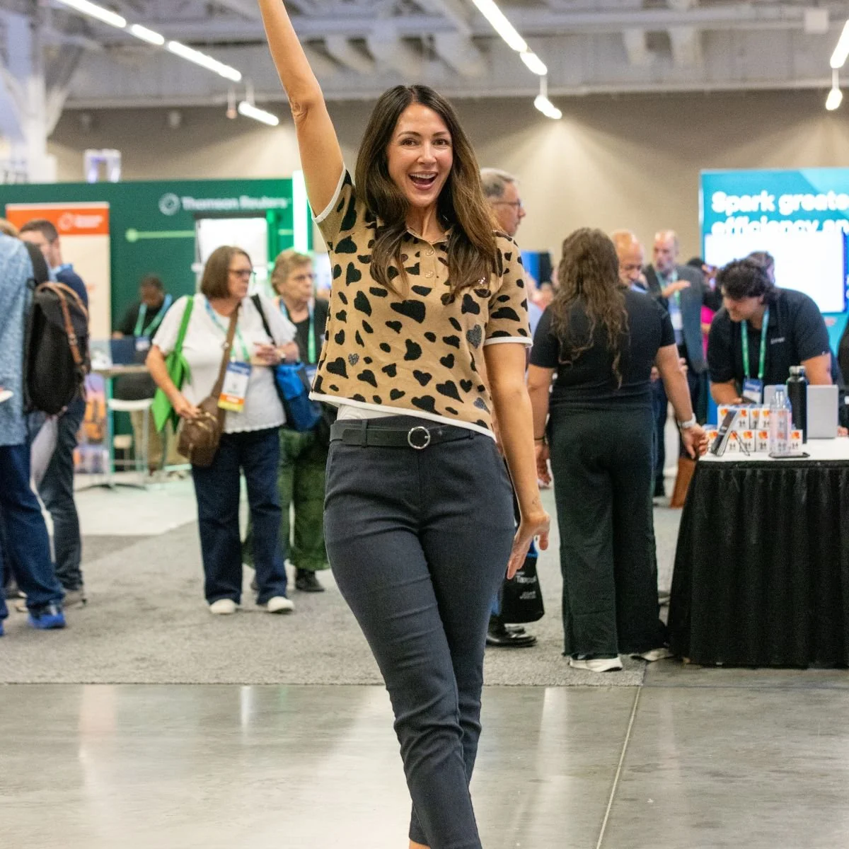 A woman with long brown hair wearing a leopard-print shirt and black pants smiling and raising her right hand at a busy indoor event or conference.