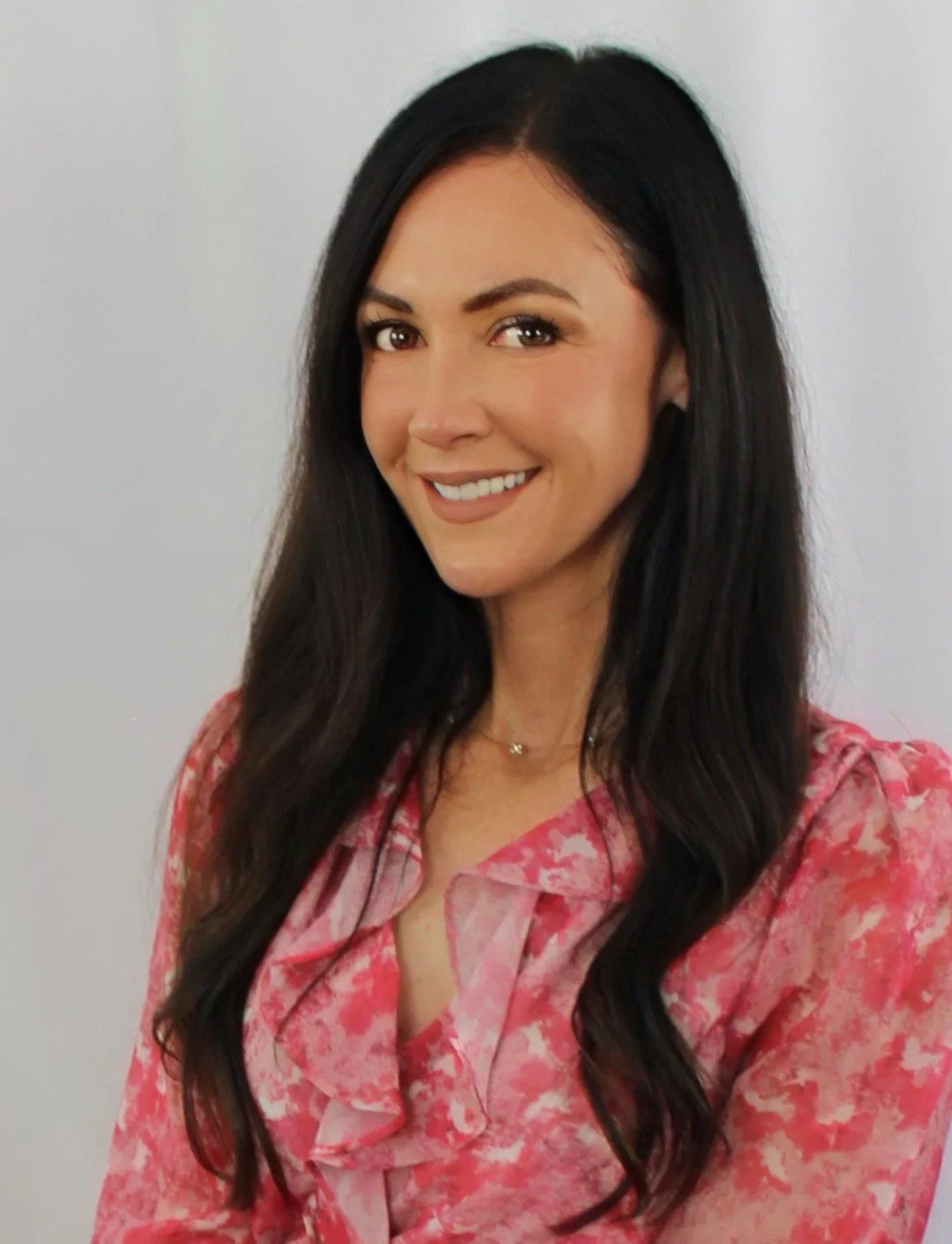 A woman with long dark hair, smiling, wearing a pink floral blouse, standing against a plain white background.