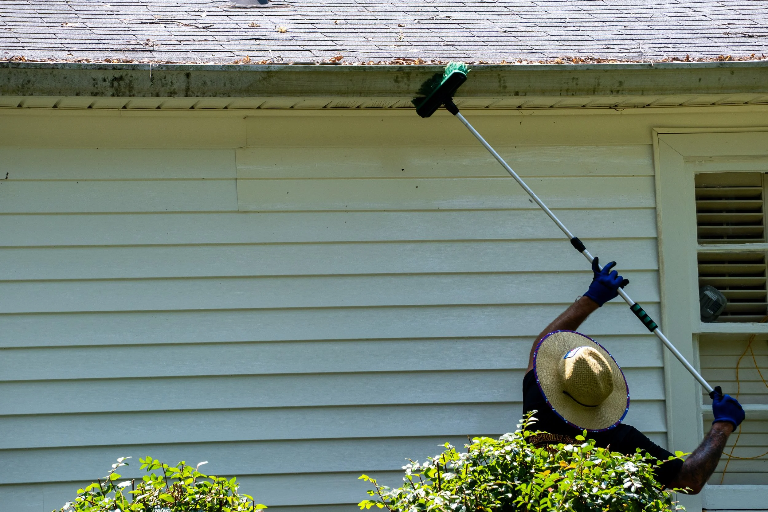 Person wearing a hat and gloves using a long-handled tool to clean or scrape the soffit of a house exterior with beige siding and a vent, with shrubbery in the foreground.
