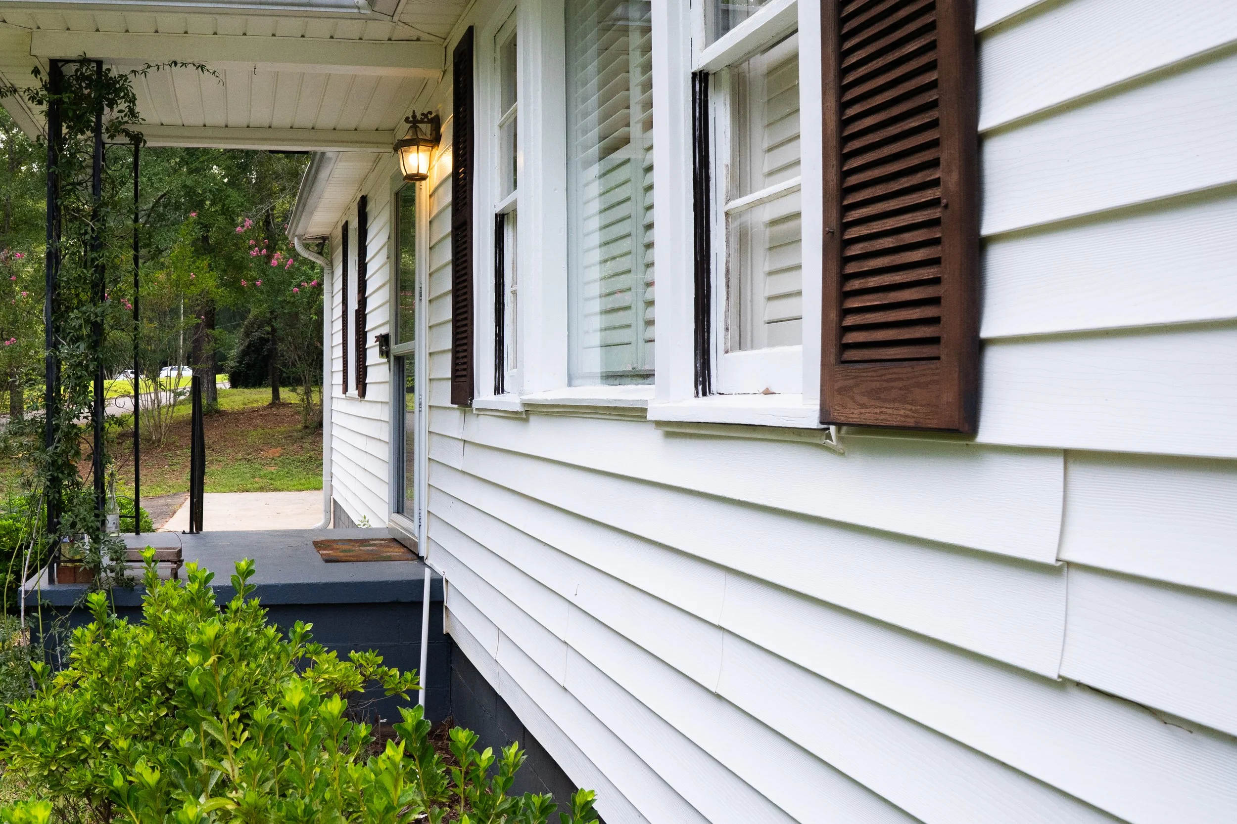 Side view of a white house with brown shutters and a porch with a light fixture, surrounded by greenery and trees.