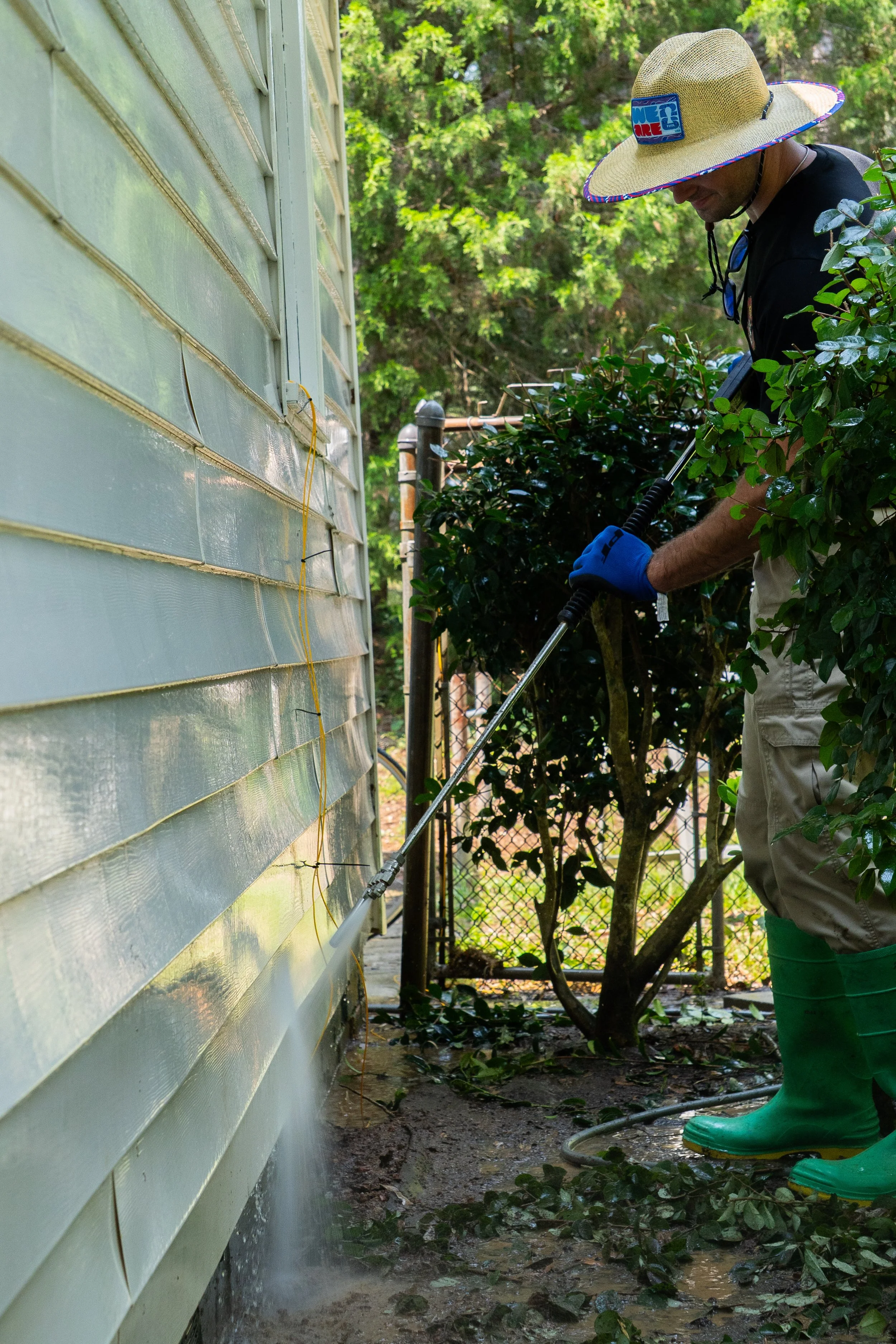 A person wearing a wide-brimmed straw hat, sunglasses, black shirt, beige cargo shorts, and bright green rain boots, watering plants outside next to a house with light-colored siding. The person uses a long-handled watering device, surrounded by gree