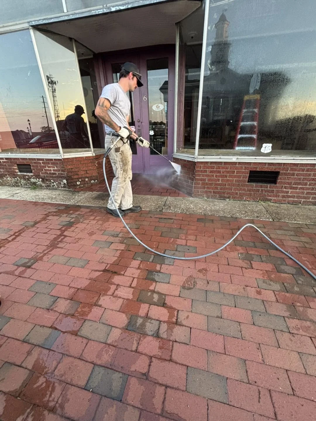 A person using a pressure washer to clean a brick sidewalk outside a building with large glass windows and a purple door.