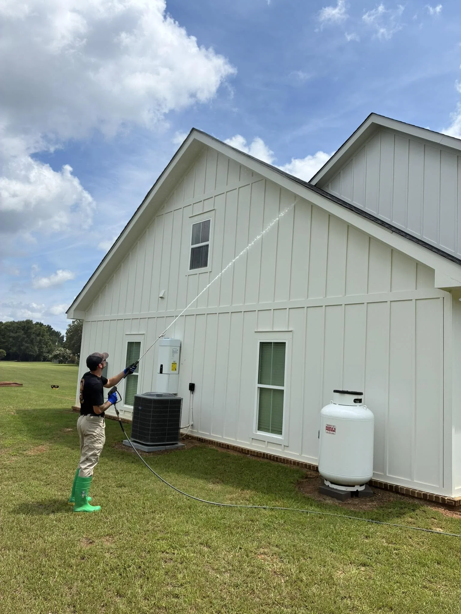 A person wearing green boots and black gloves using a pressure washer to clean the side of a white house with grass in the yard and blue sky with clouds overhead.