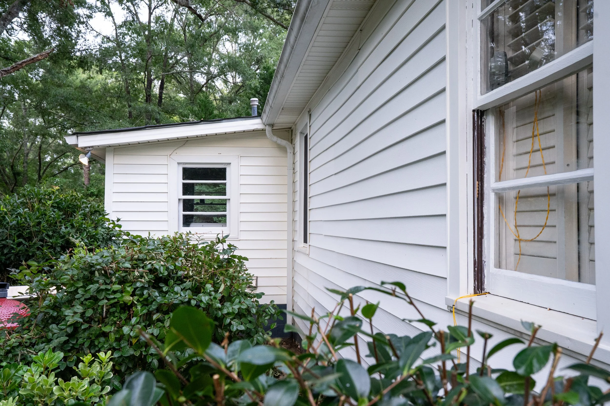 Side view of a house with white vinyl siding, several windows, and a lush green shrub in the foreground. Trees are visible in the background under an overcast sky.