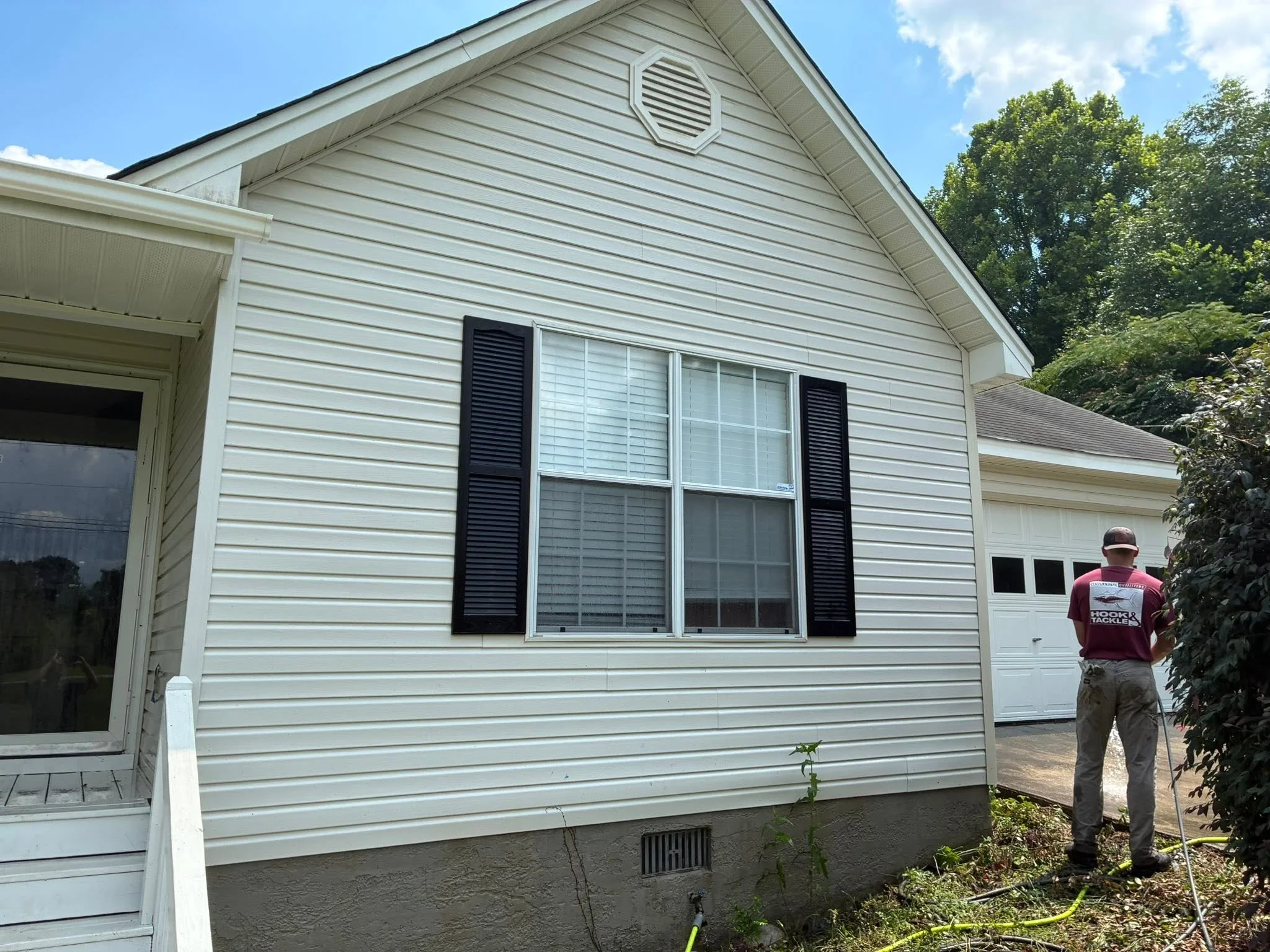 The back of a two-story house with white siding and a large window with black shutters. The house has a gable roof, an exhaust vent, and a small ladder on the side. Two people are working outside near the house, one wearing a maroon shirt with a logo