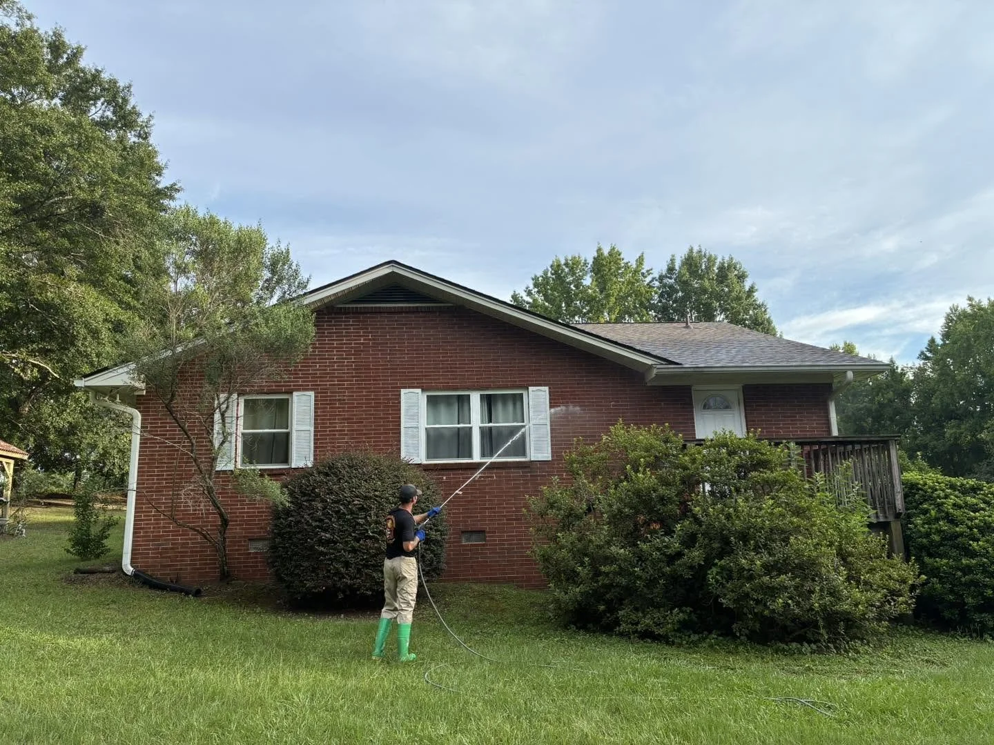 A person wearing green rain boots pressure washing the side of a red brick house, with trees and bushes around it, under a blue sky with some clouds.
