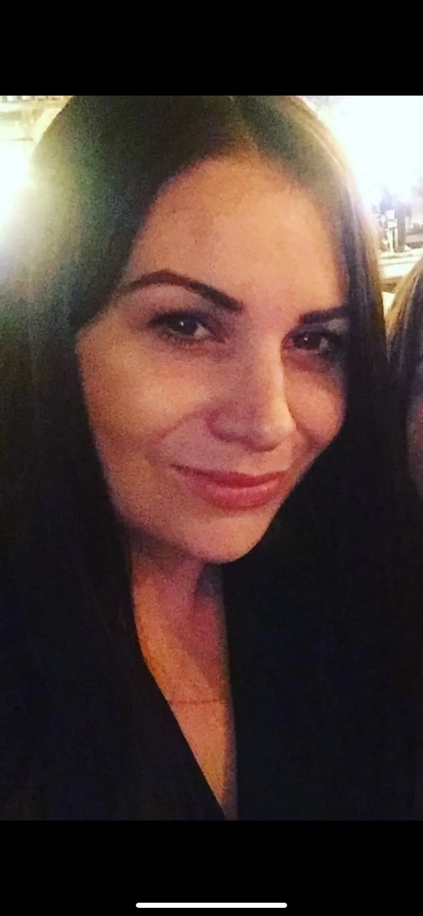 Close-up of a woman smiling in a dimly lit setting, with dark hair and makeup.