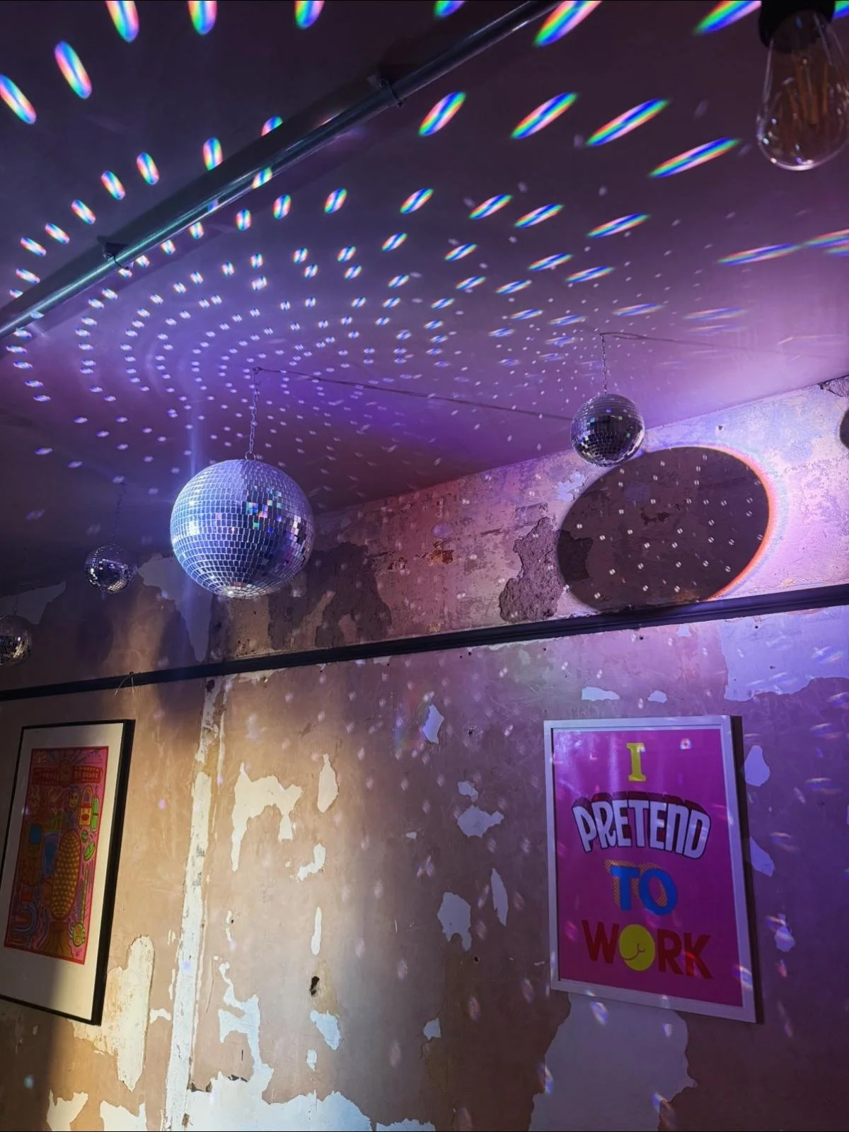 Disco balls hanging from the ceiling create light reflections on the dark ceiling, with one prominent disco ball casting a shadow on a wall with peeling paint. The wall features colorful framed artwork and a pink poster reading 'I PRETEND TO WORK'.