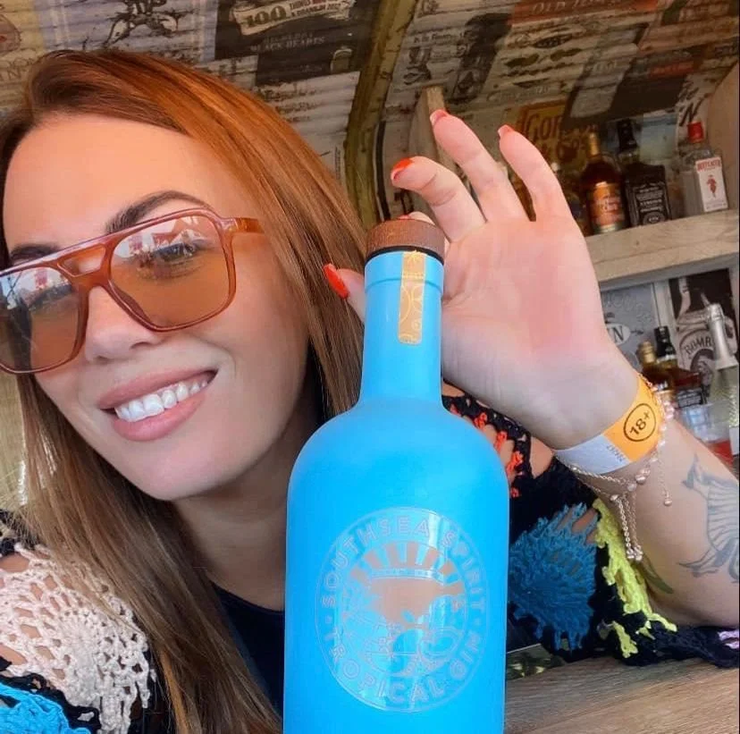 A woman with red hair and sunglasses takes a selfie in a bar or pub, holding a blue bottle in front of her.