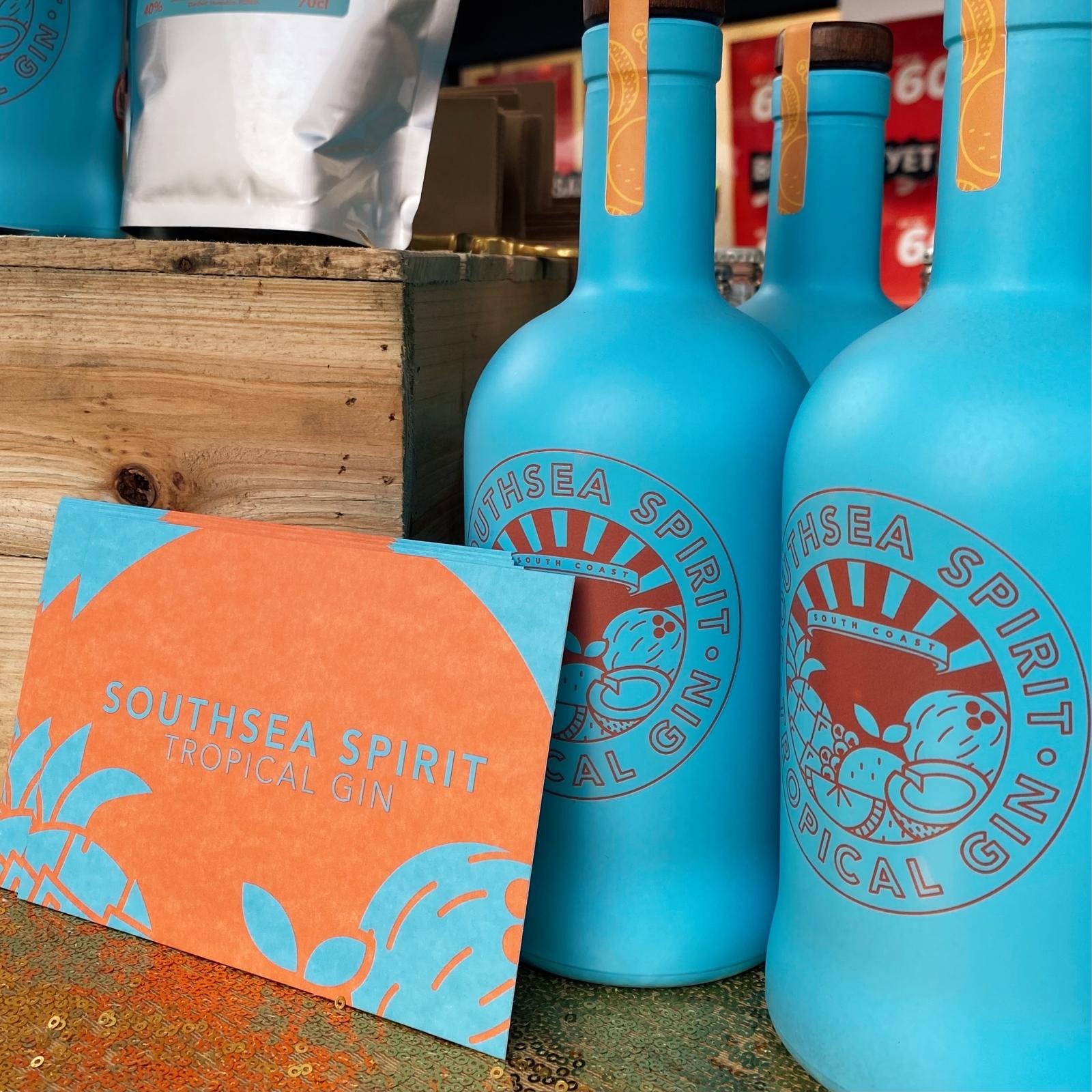 Blue bottles with a logo reading 'Southsea Spirit' and a graphic of citrus fruits and a cocktail glass. An orange card with similar branding and text 'Southsea Spirit Tropical Gin' is in the foreground, set on a wooden surface.