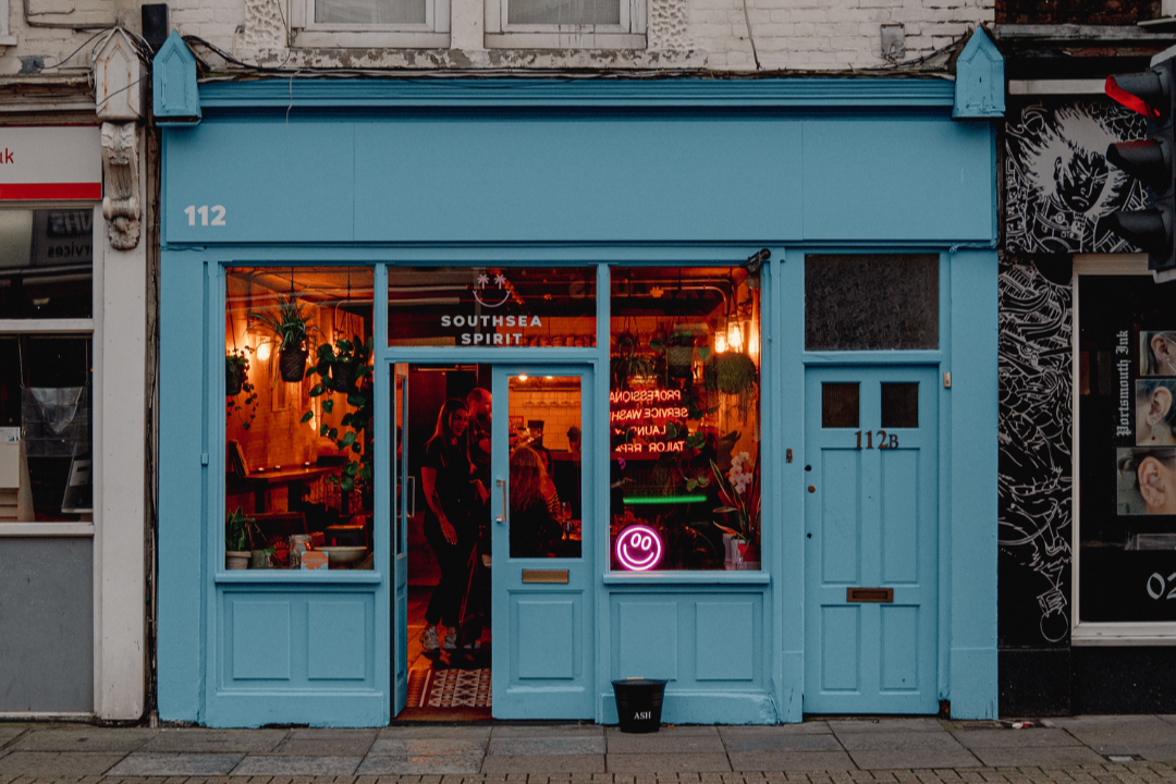 A blue storefront with the number 112 on the upper left corner, decorated with small blue triangular roof details. Inside, warm lighting and green plants are visible through large glass windows. Neon signs and people are also visible inside.