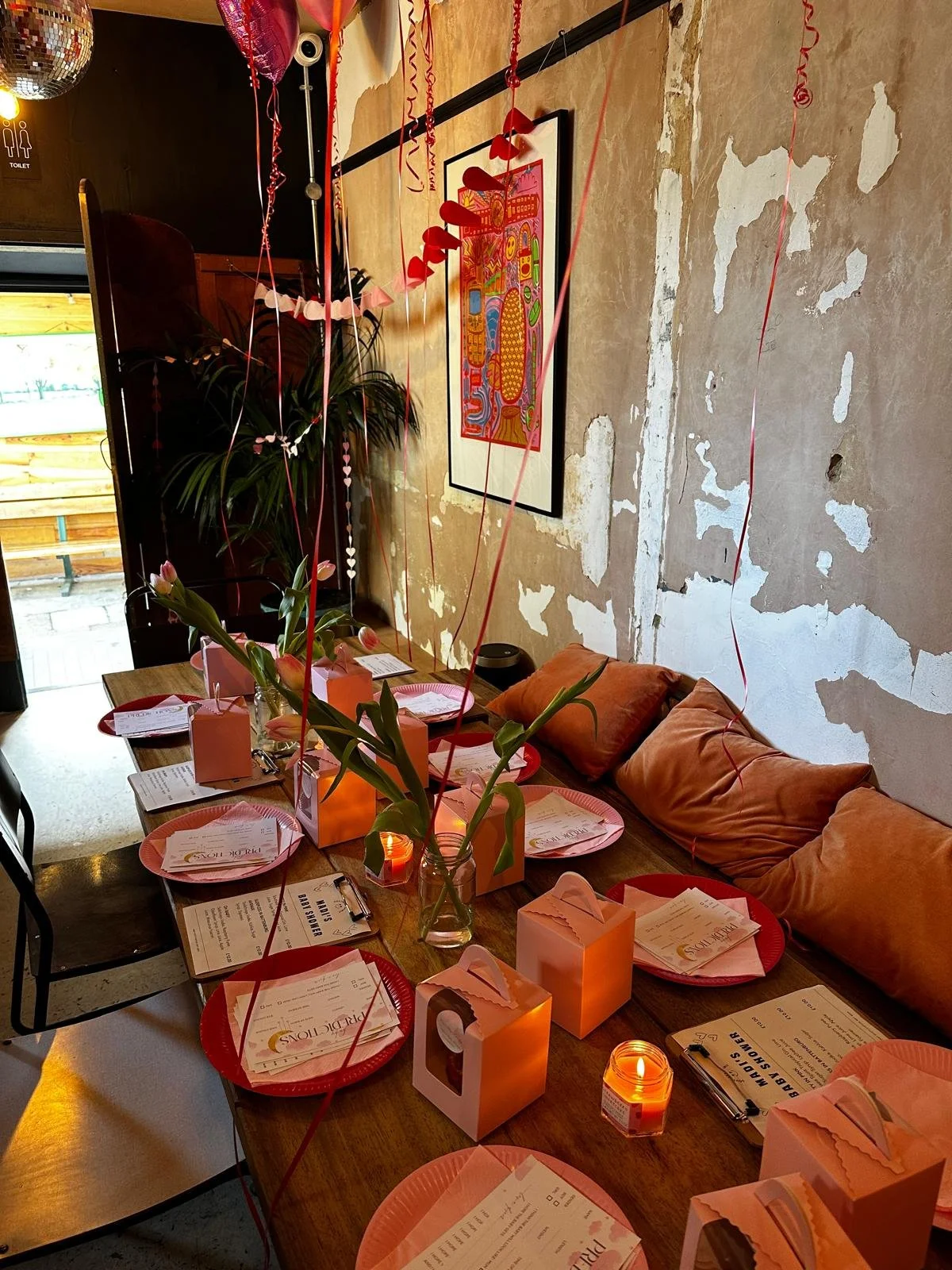 A decorated table set for a celebration, featuring pink plates, menu cards, pink gift boxes, lit candles, and floral arrangements with pink lilies. Balloons and red ribbons hang from the ceiling, and there is a colorful artwork on the wall in the background. The setting appears cozy and festive.
