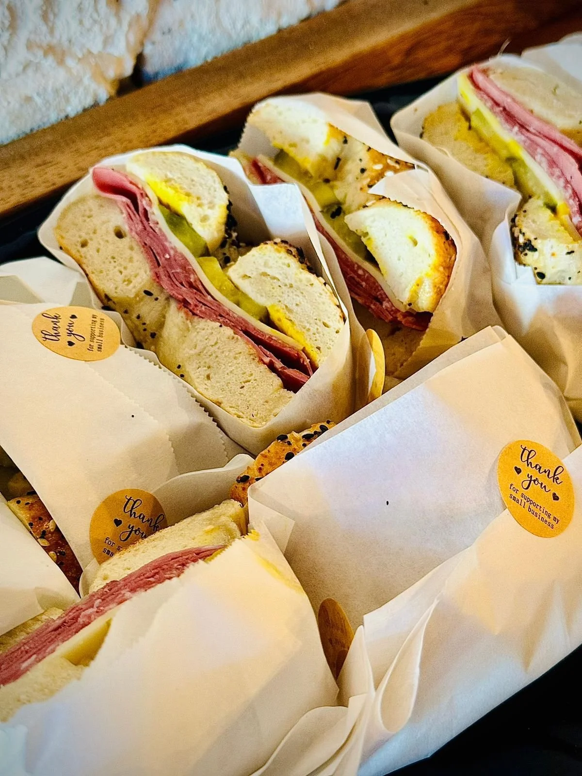 Several sandwiches wrapped in white paper with yellow stickers that say 'thank you for supporting my small business.' The sandwiches contain ham, cheese, and pickles, and are served in white bread.