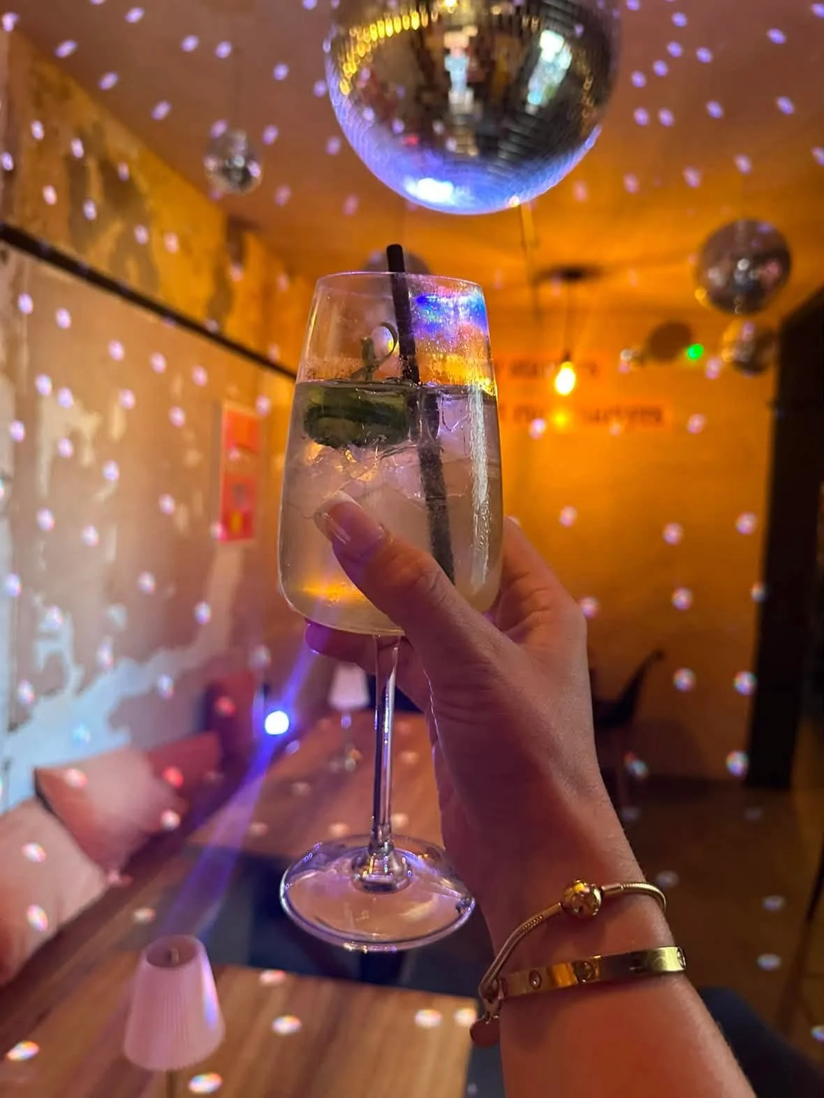 A person holding a glass of white wine with ice, lime, and a black straw, inside a dimly lit bar or club with a disco ball and colorful lights reflecting on the walls.