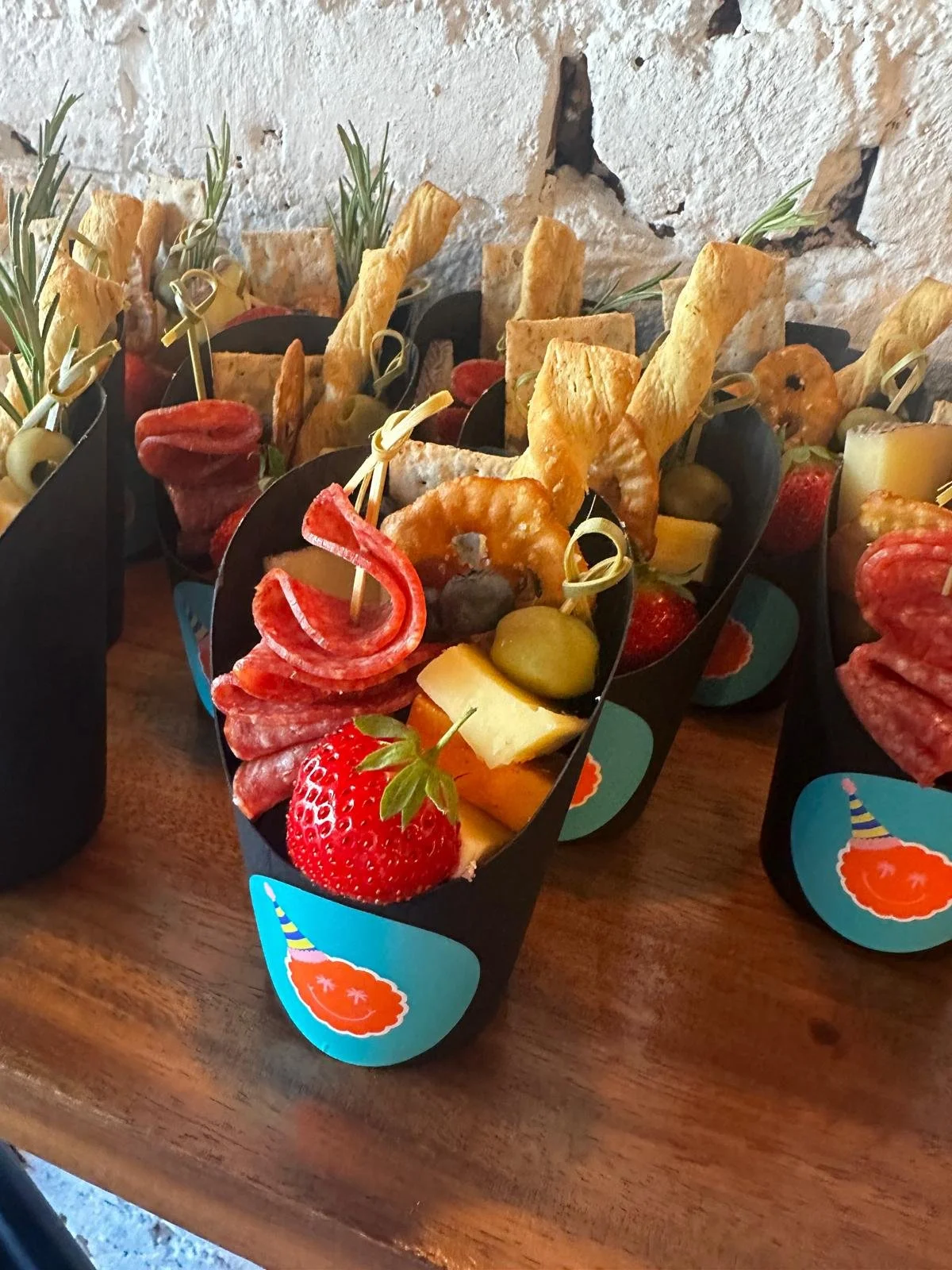 Party favor boxes filled with cured meats, cheese, strawberries, grapes, crackers, and herbs, placed on a wooden surface.