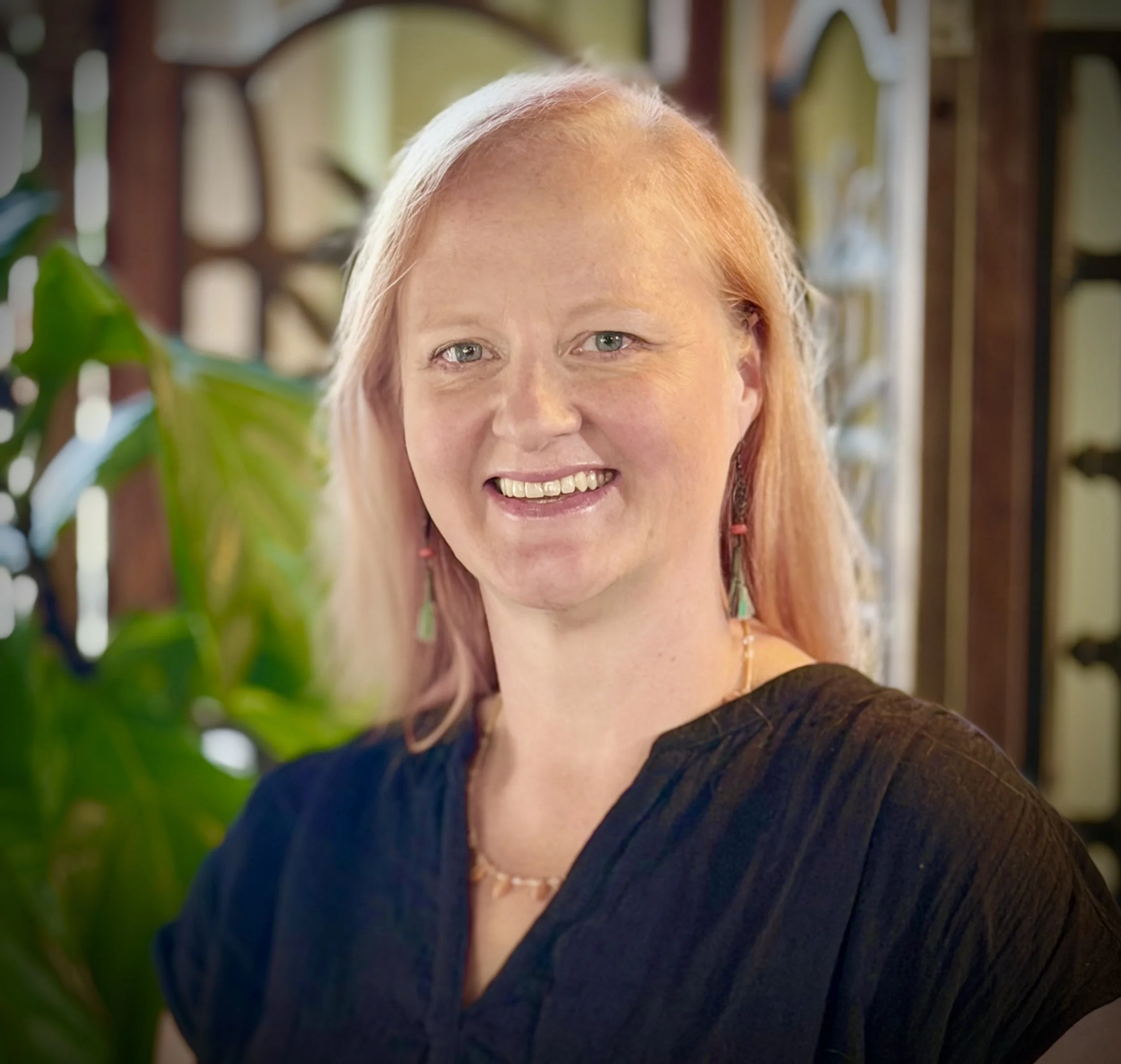 Close-up of a woman's face smiling, with blonde hair and green eyes, with a blurred background and some green plants in the foreground.