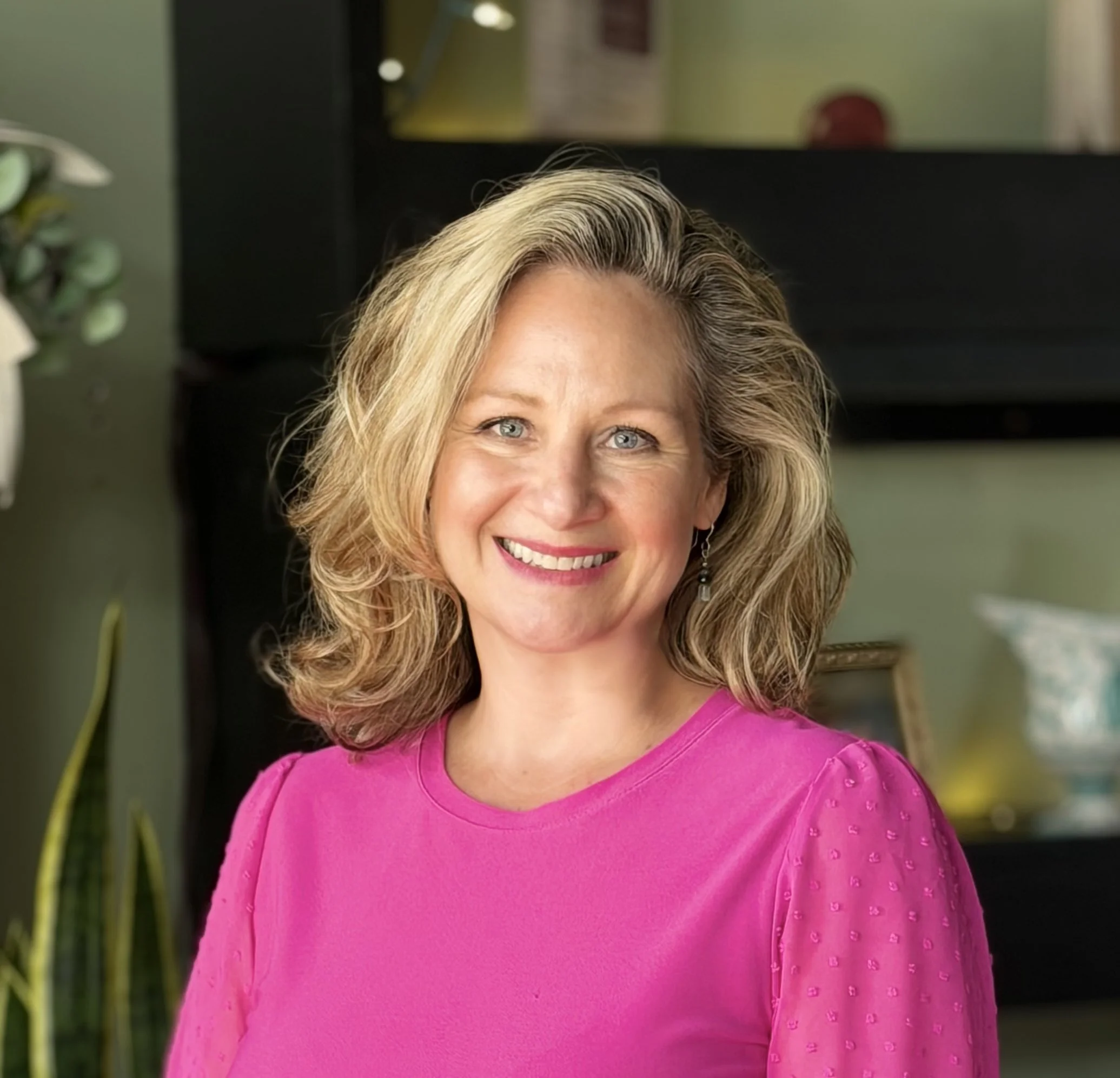 A woman with blonde hair smiling, wearing a pink top with textured detailing, standing indoors with green walls, a plant on the left, and a mirror or glass behind her.