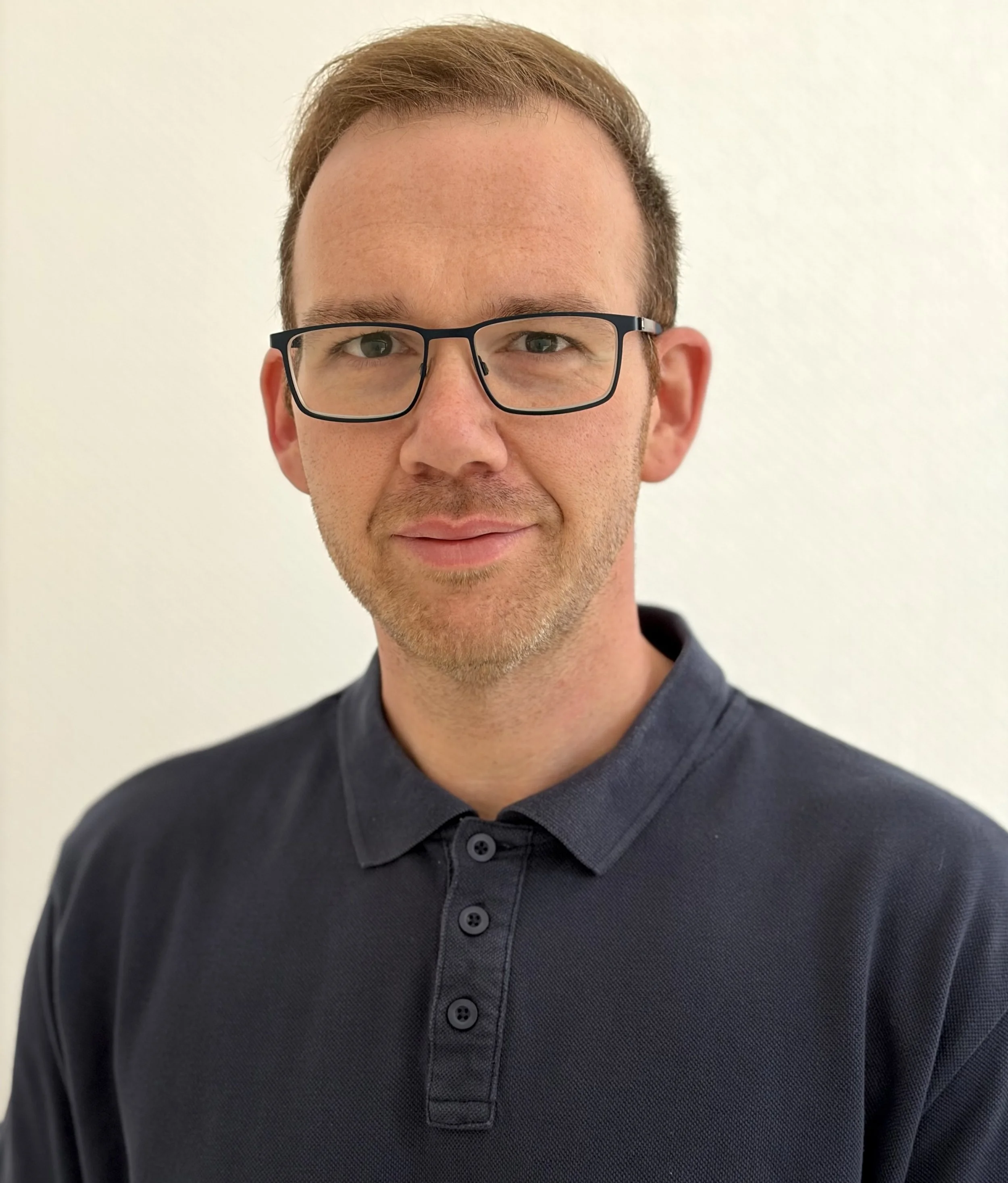 Nahaufnahme eines Mannes mit braunen Haaren, Brille und dunklem Hemd vor einem weißen Hintergrund.