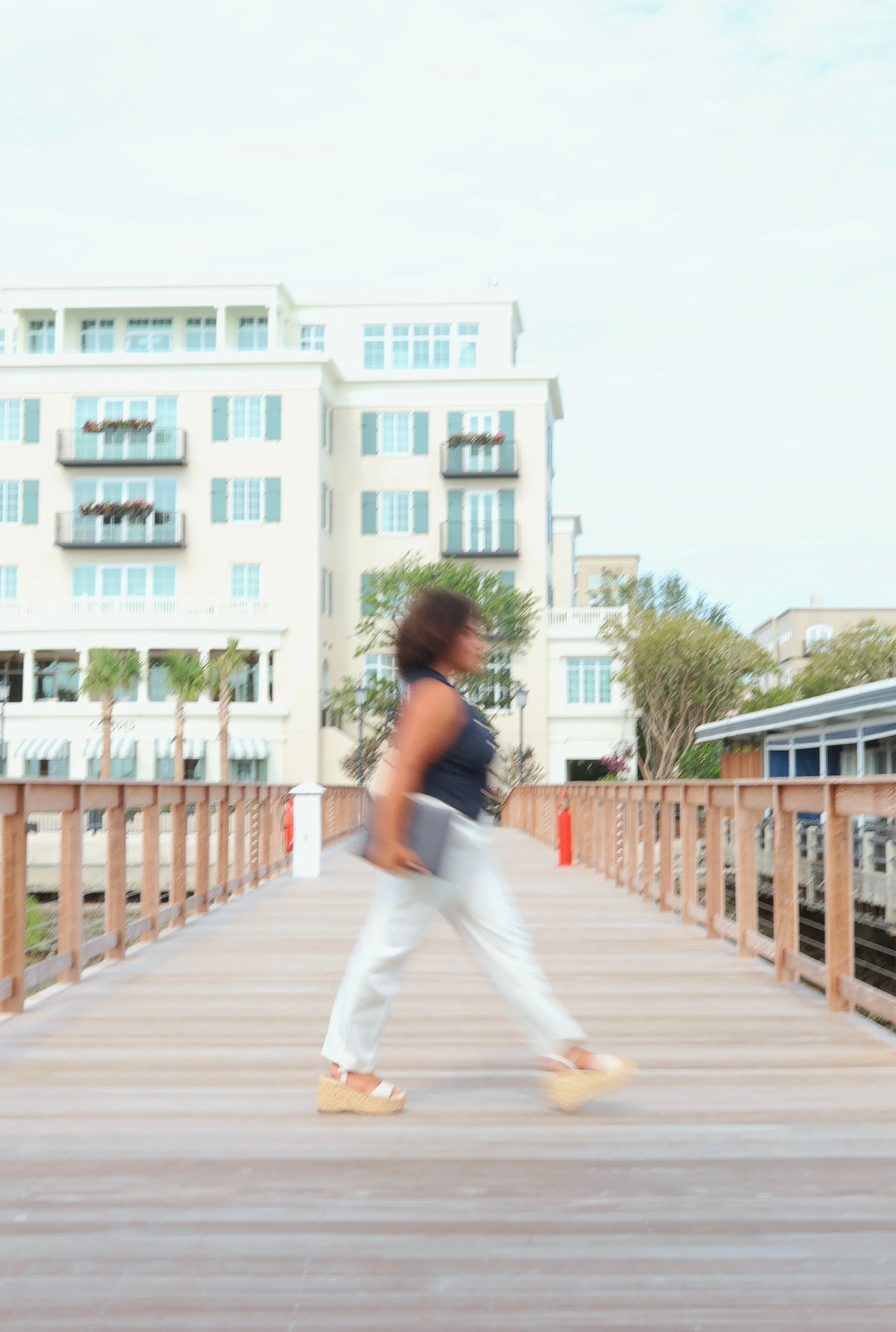 Woman walking on a wooden bridge in front of a white multi-story building with blue shutters and balconies with flowers, under a partly cloudy sky.