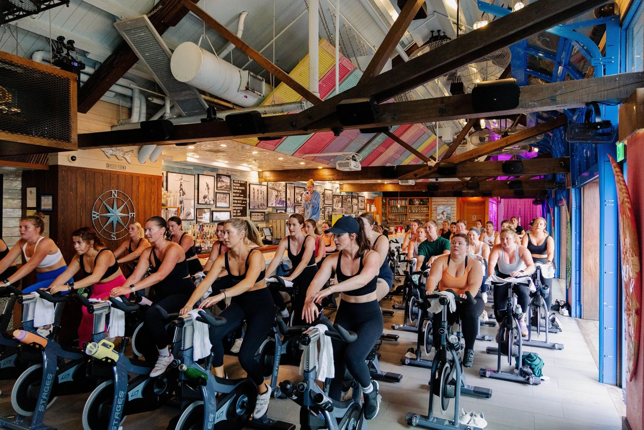 Group of women participating in an indoor spin class on stationary bikes, with colorful wall decor and a woman leading the class in the background.