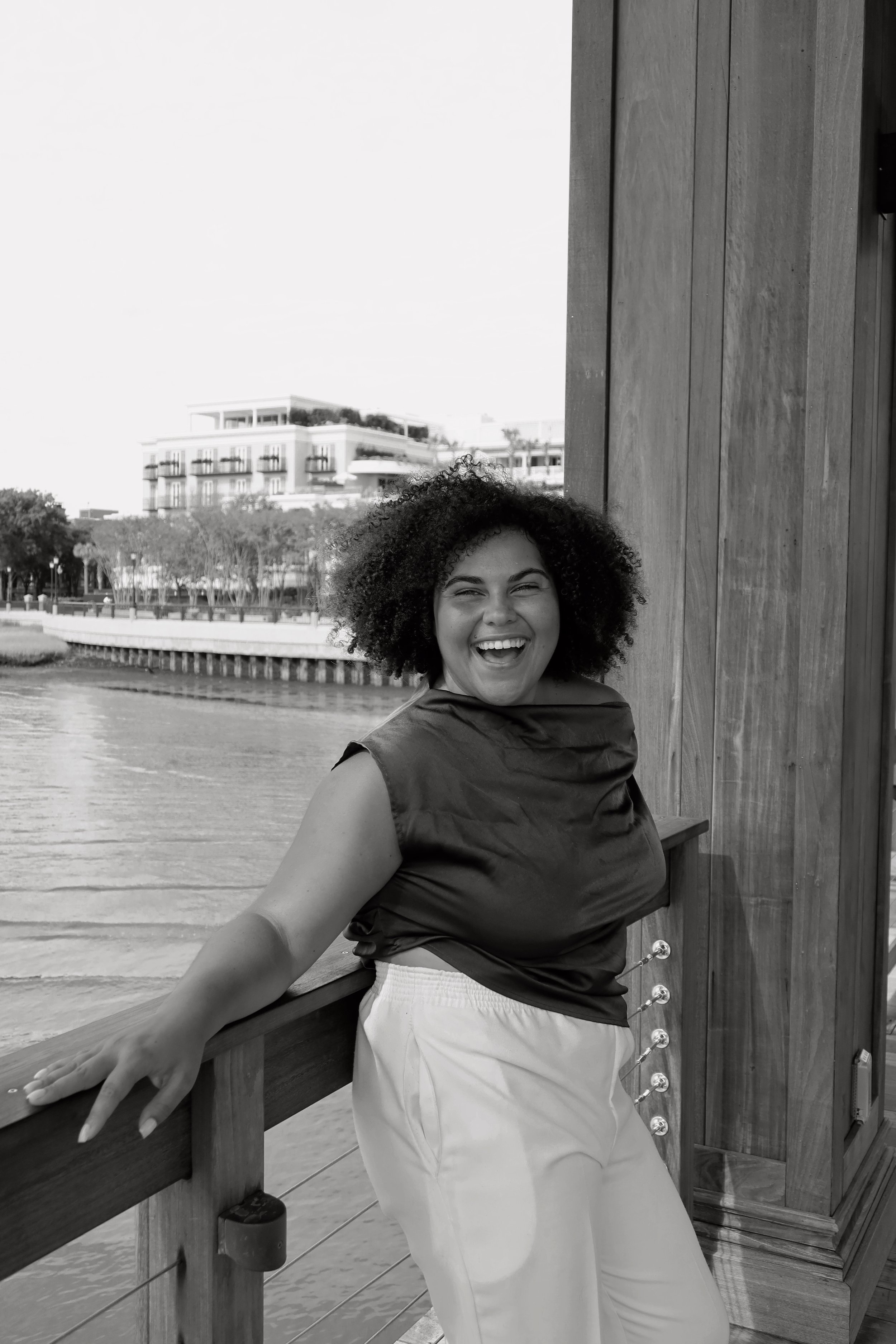 A joyful woman with curly hair standing outdoors by a wooden railing, smiling widely.