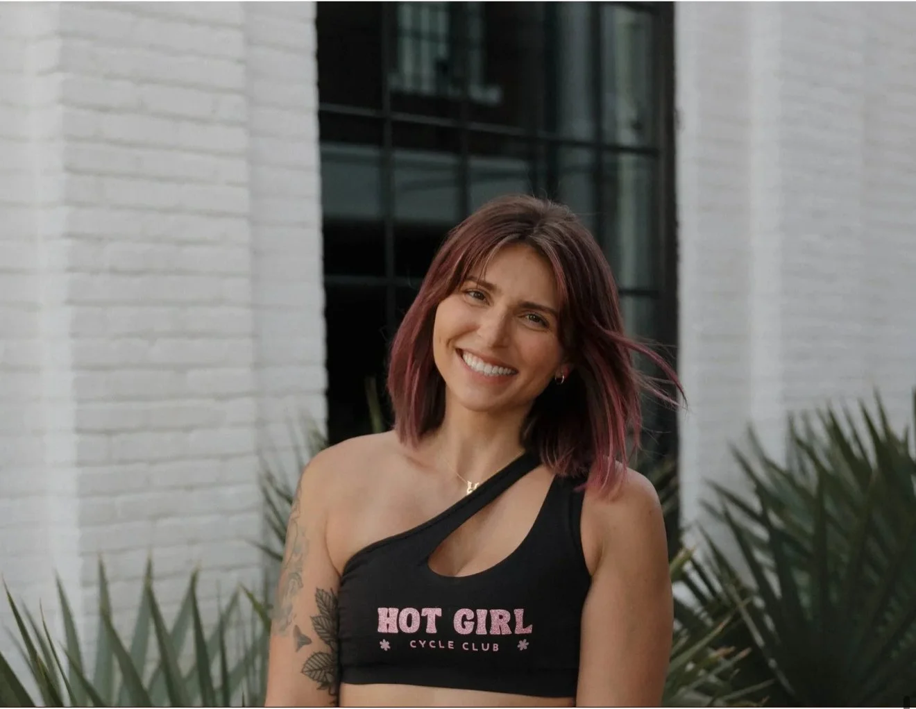 A young woman with shoulder-length purple hair, smiling, wearing a black sports bra with pink text that reads 'Hot Girl Cycle Club,' standing outdoors in front of a white brick wall and green plants.