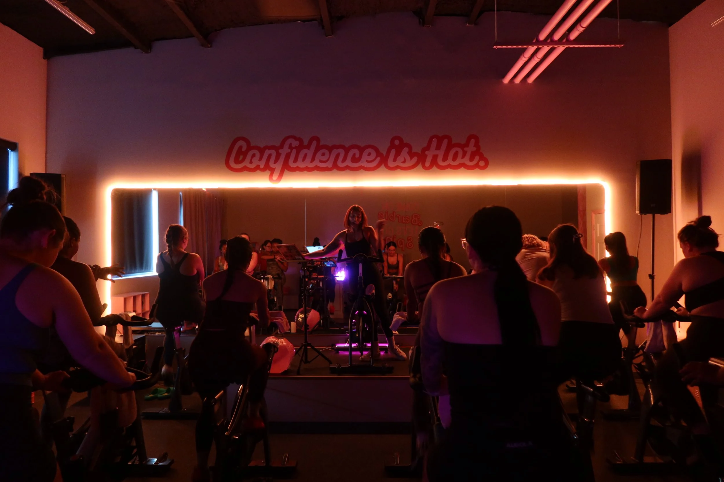 Group fitness class in a dimly lit studio with a mirror, neon lighting, and a sign that reads "Confidence is Hot." Participants are on stationary bikes, and an instructor is leading the session.