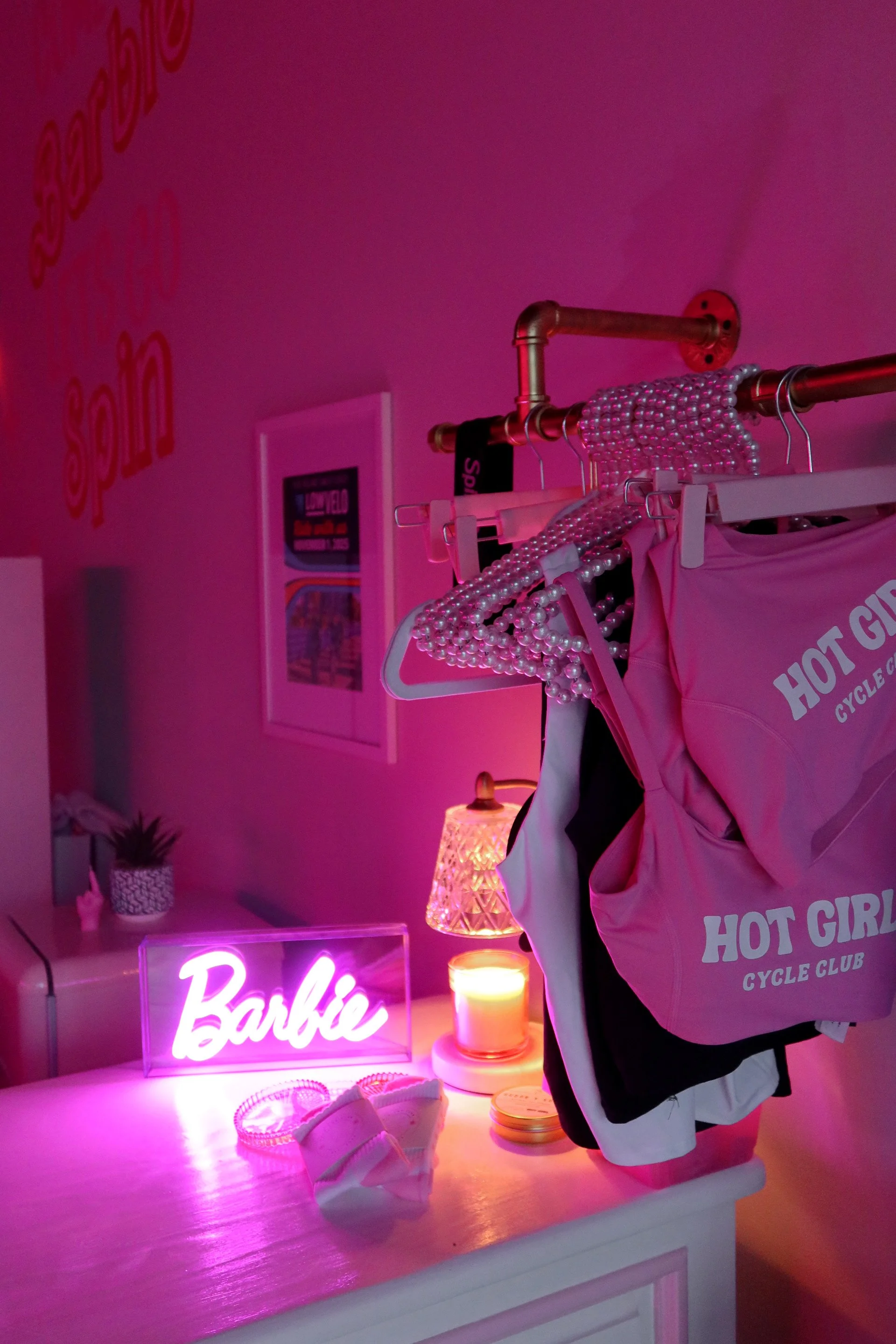 A pink room decorated with neon signs, including a "Barbie" sign, and a clothing rack with pearl necklaces and clothing. A small table with a candle and a potted plant, with a soft pink lighting creating a cozy atmosphere.
