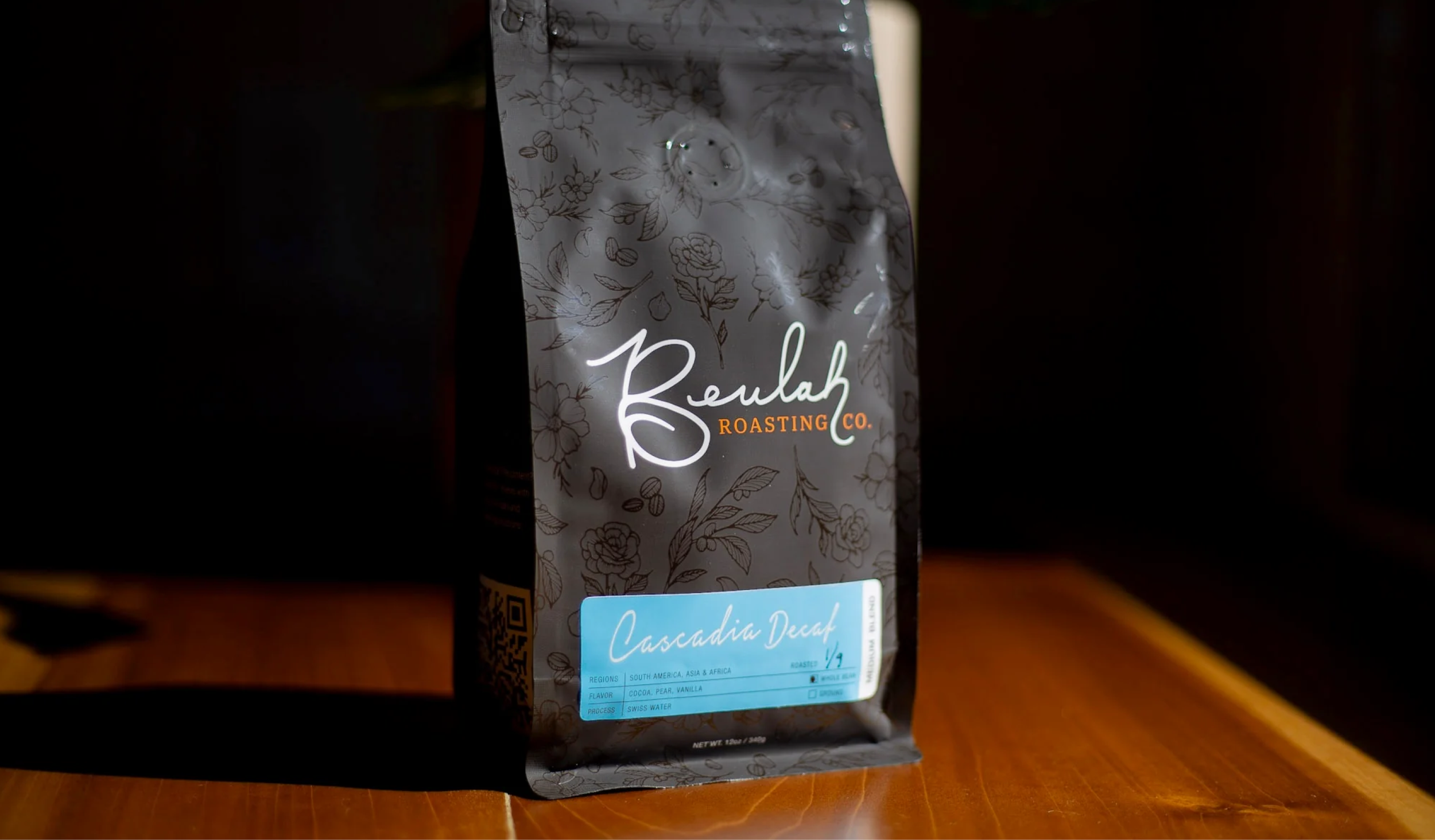 Packaging of Beulah Roasting Co. Cascadia Decaf coffee with floral design, a blue label indicating the coffee's name, origin, flavor, and process, placed on a wooden surface.