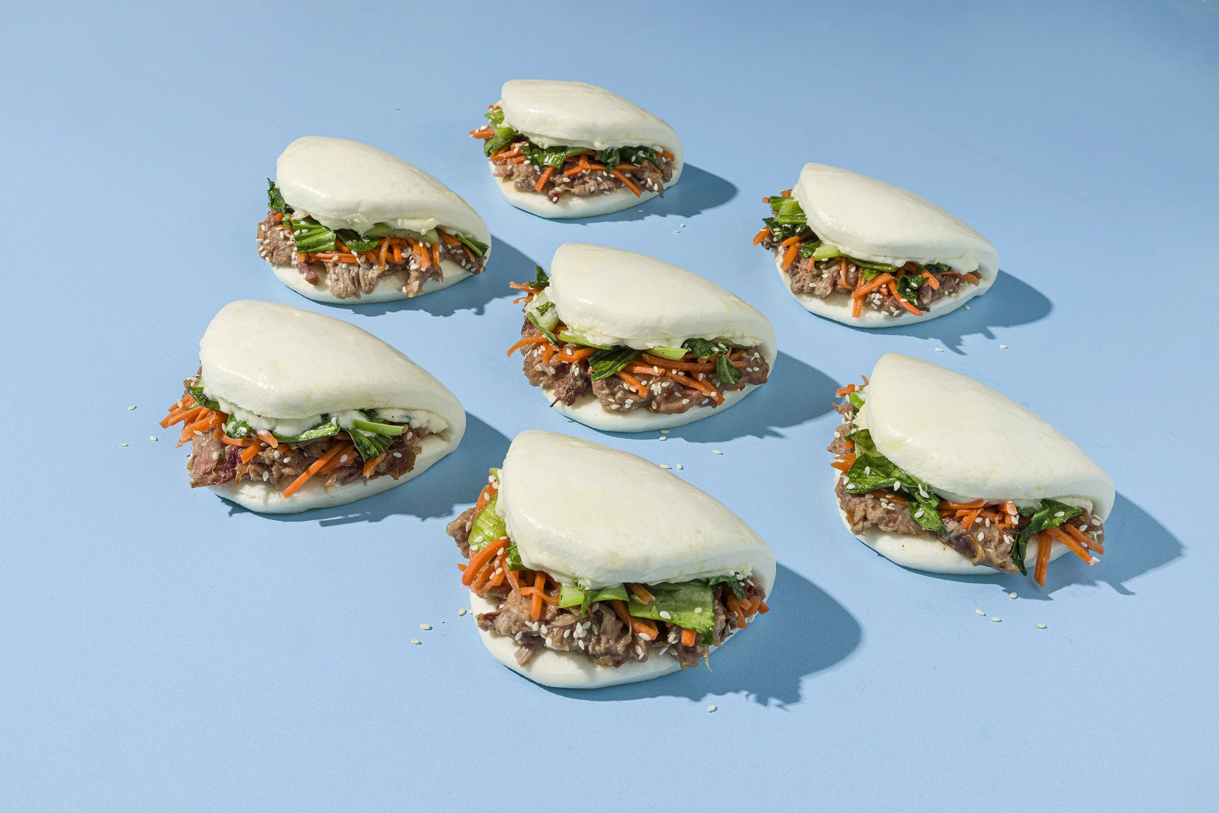 Several steamed bao buns filled with shredded carrots, greens, and minced meat on a light blue background.