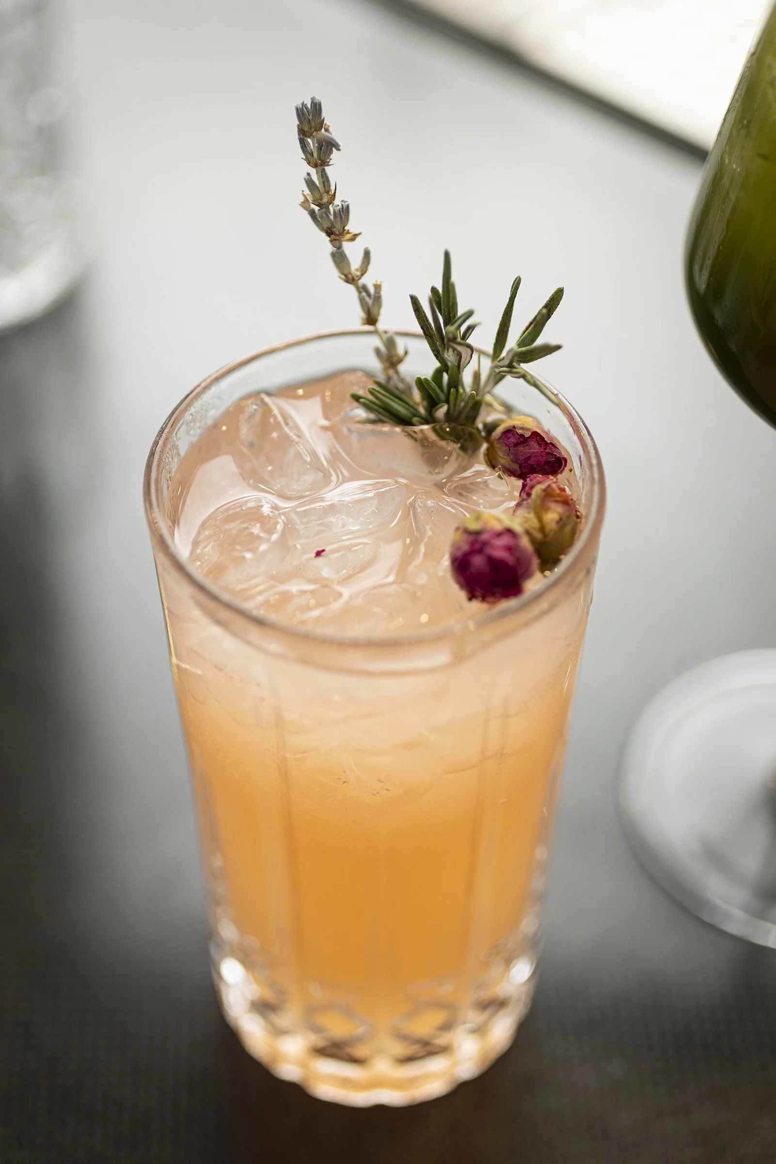 A tall glass with a pink drink, ice cubes, garnished with sprigs of lavender and rosemary, and small dried rosebuds.