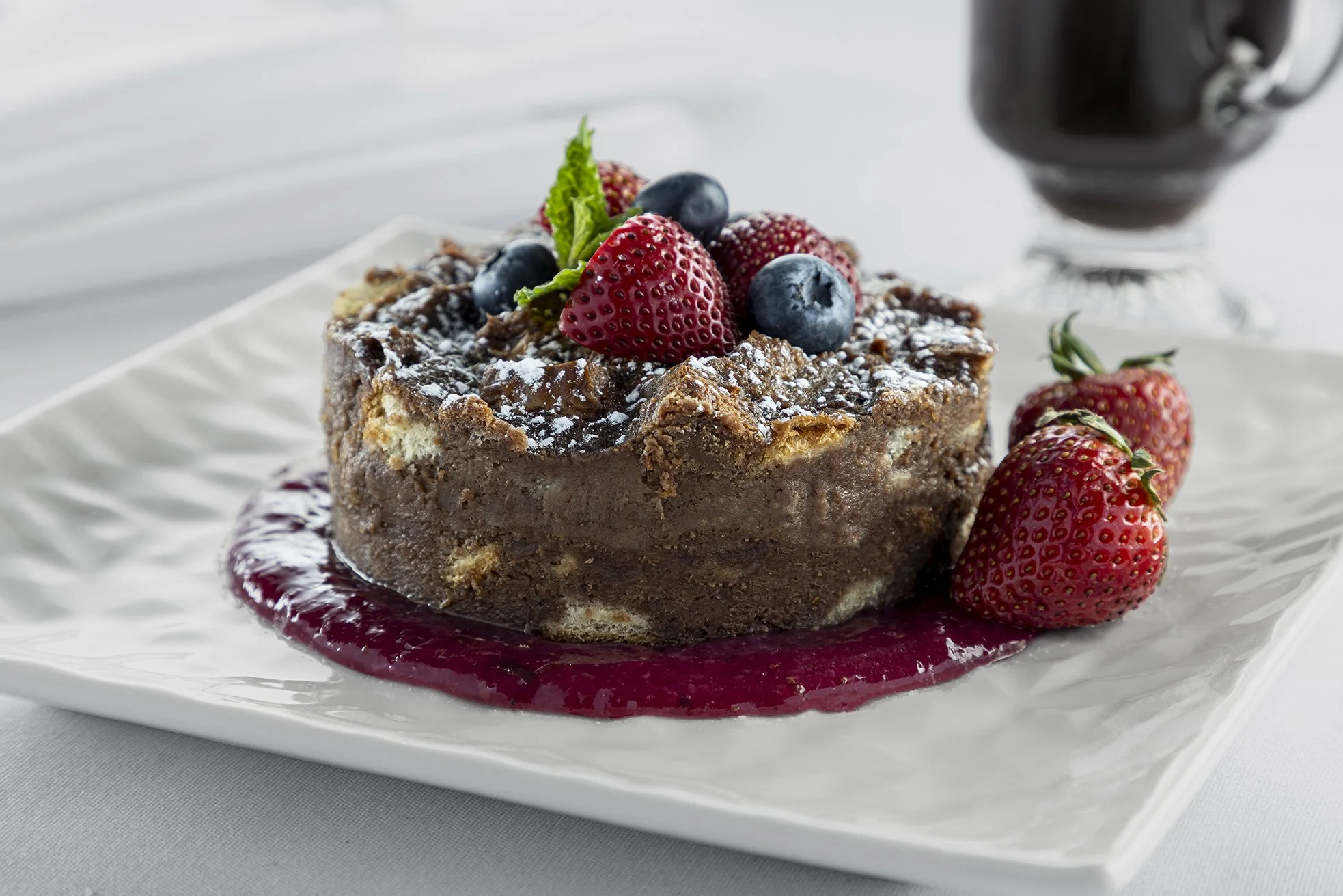 Chocolate cake topped with strawberries, blueberries, and a mint leaf, with berry sauce drizzled on a white plate.