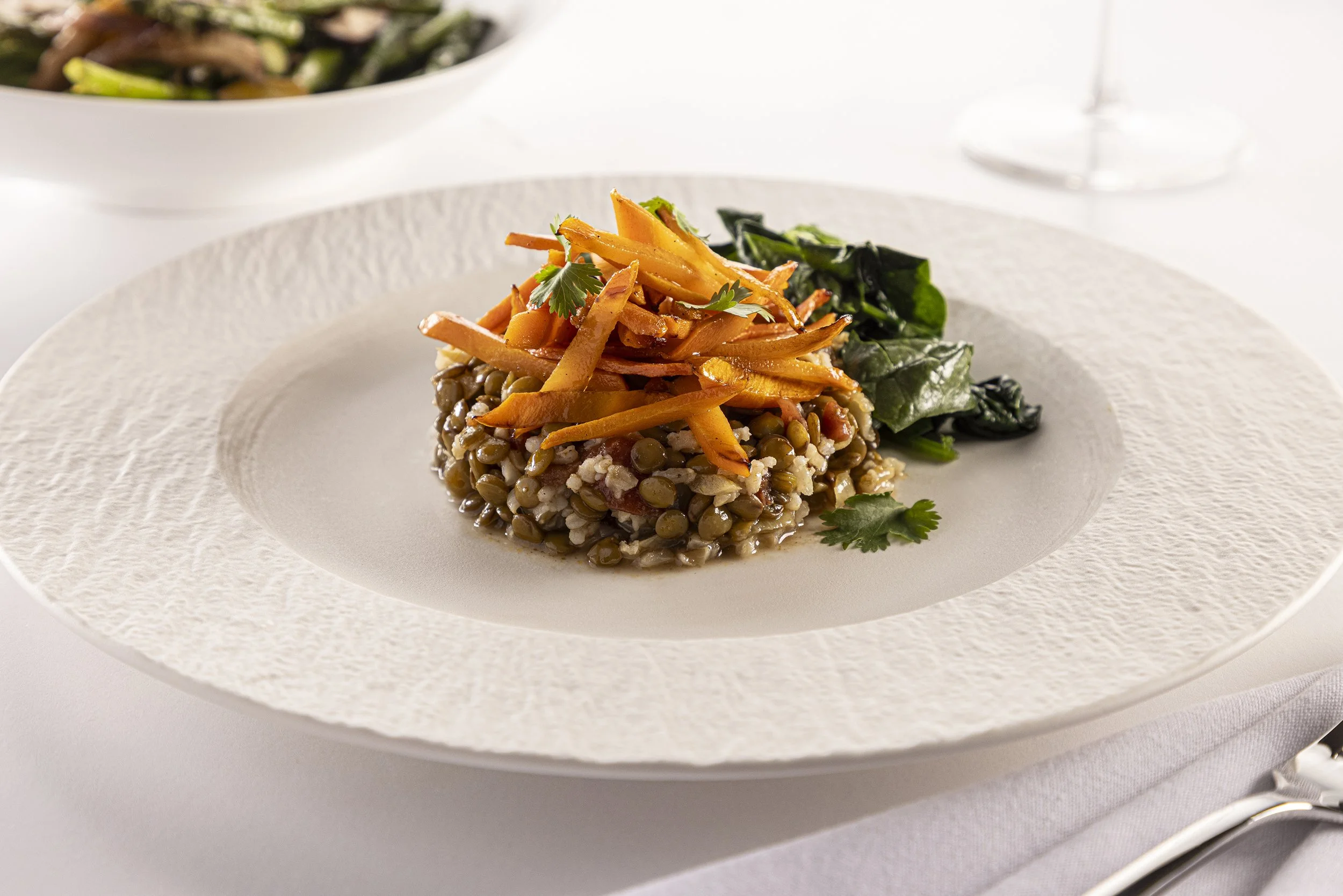 A plated dish containing lentils topped with julienned carrots and garnished with parsley, served with a side of sautéed greens, on a textured white plate.