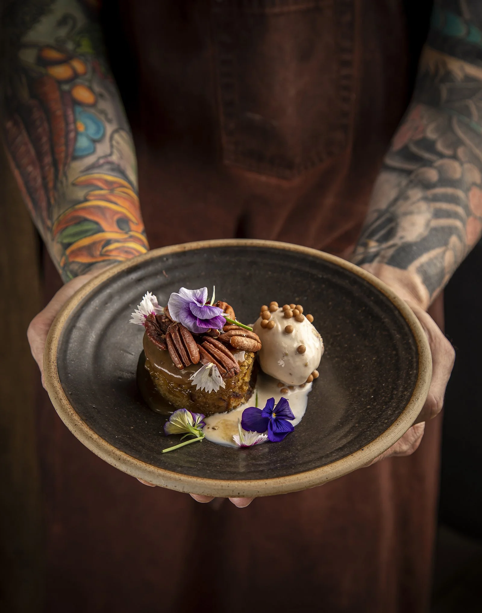A person with colorful tattooed arms holding a dark ceramic bowl with a dessert that includes a cake, ice cream, and edible flowers.