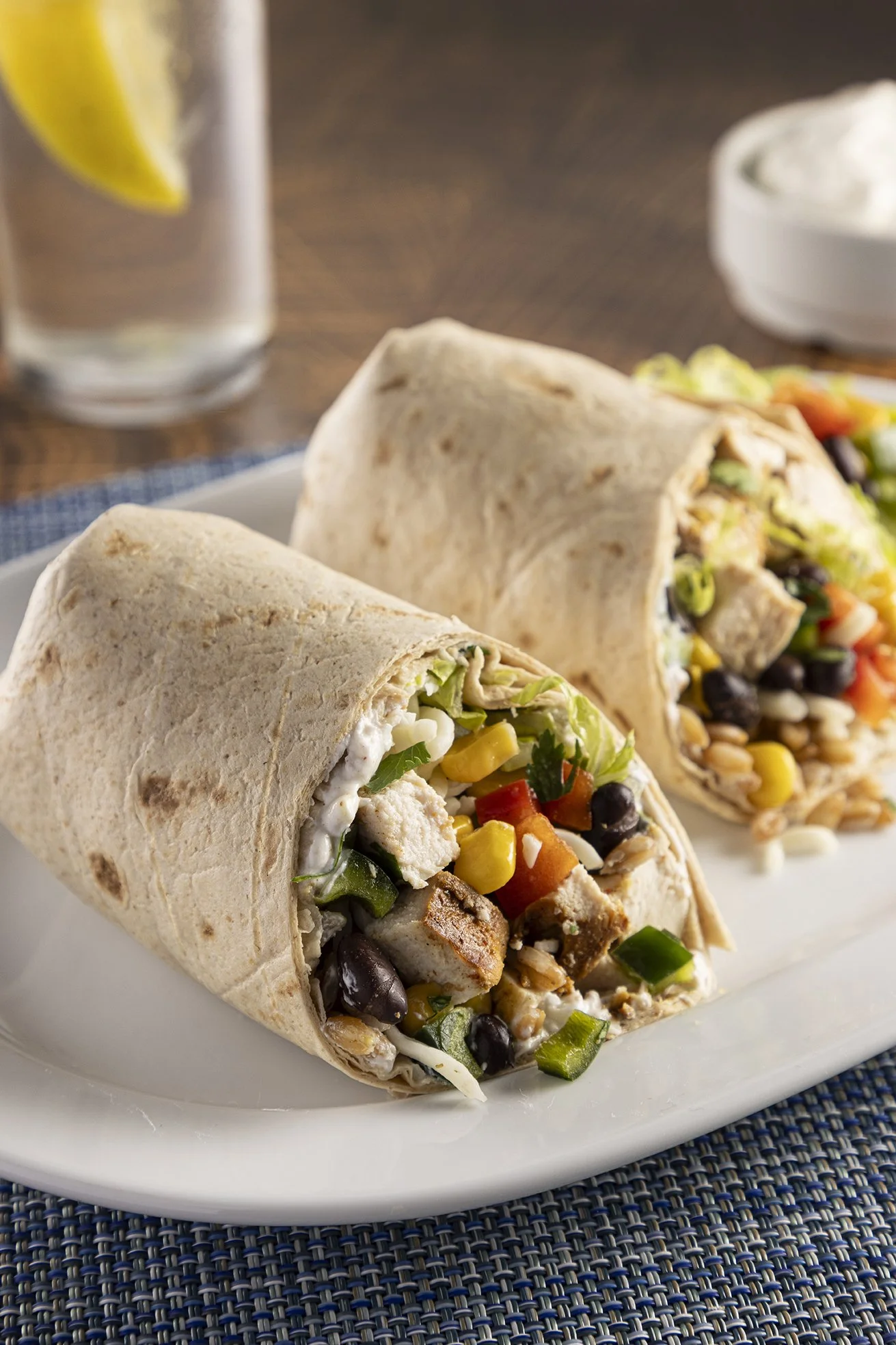 Two chicken burritos stuffed with vegetables, beans, and cheese on a white plate, with a glass of lemon water and a bowl of sour cream or dip in the background.