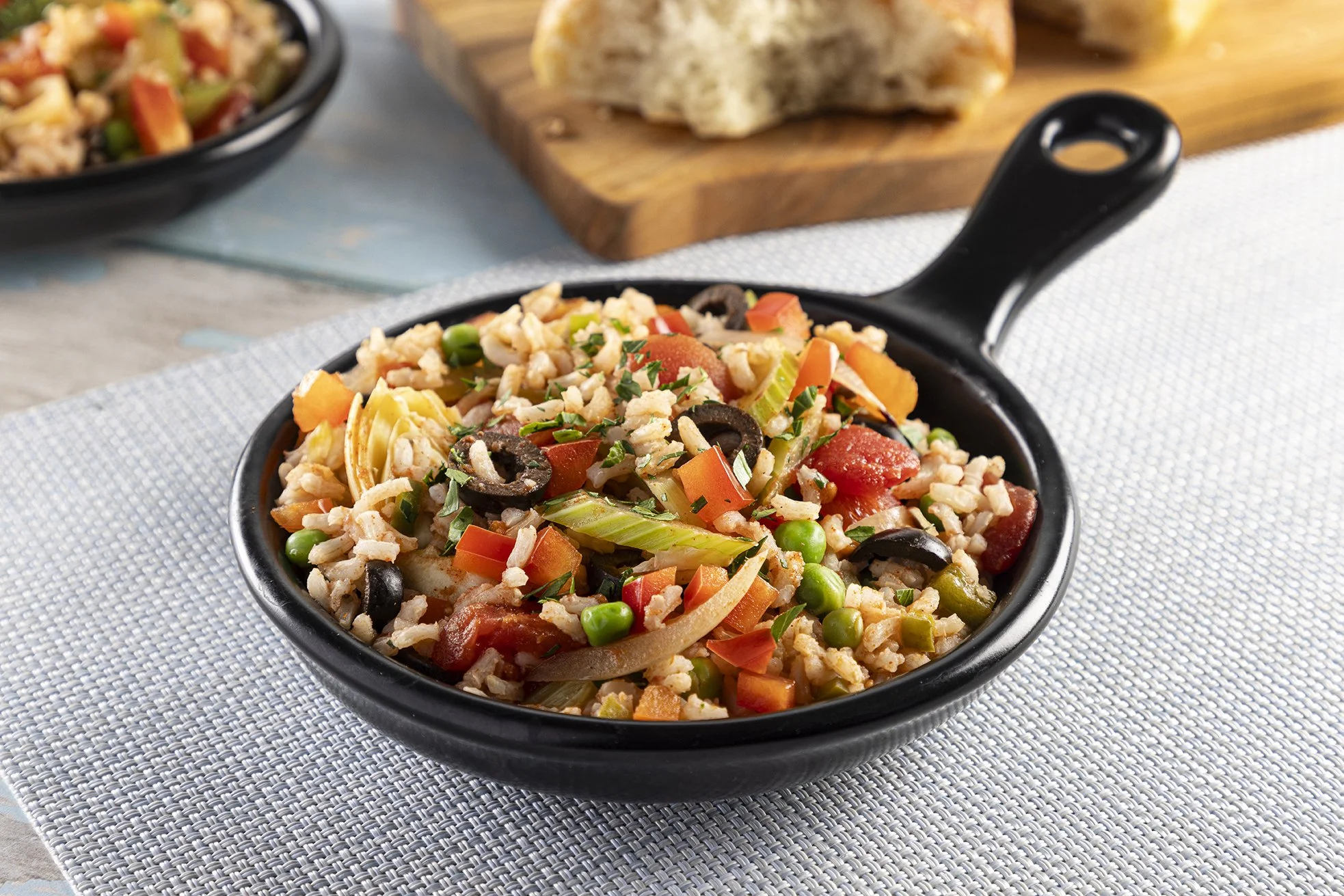 A black bowl of rice with vegetables and black olives on a gray textured placemat.
