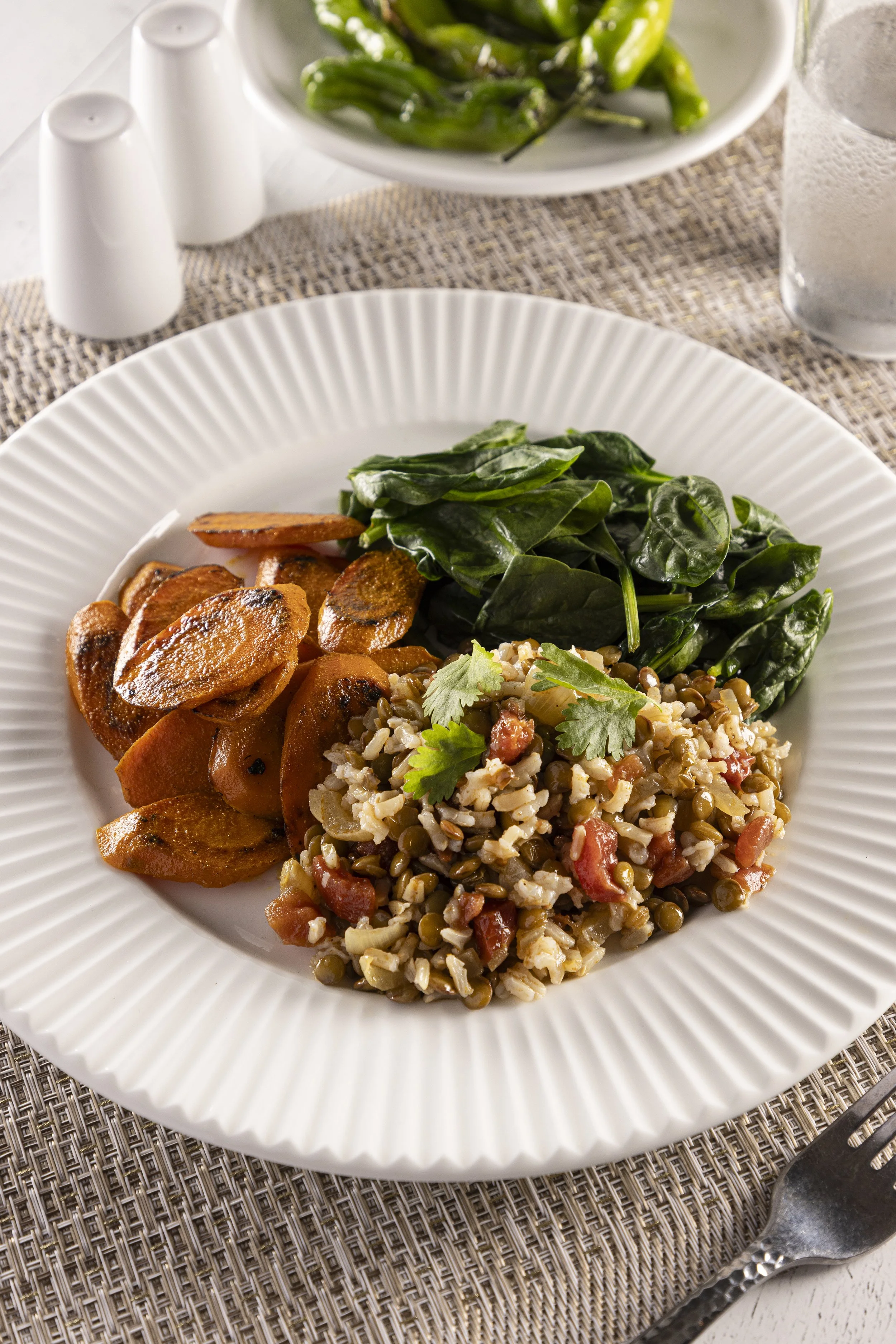 A white plate with sautéed sweet potatoes, cooked spinach, and a bed of lentil and rice mixture garnished with cilantro. In the background, a bowl of cooked spinach, salt, pepper shakers, and a glass of water on a woven placemat.