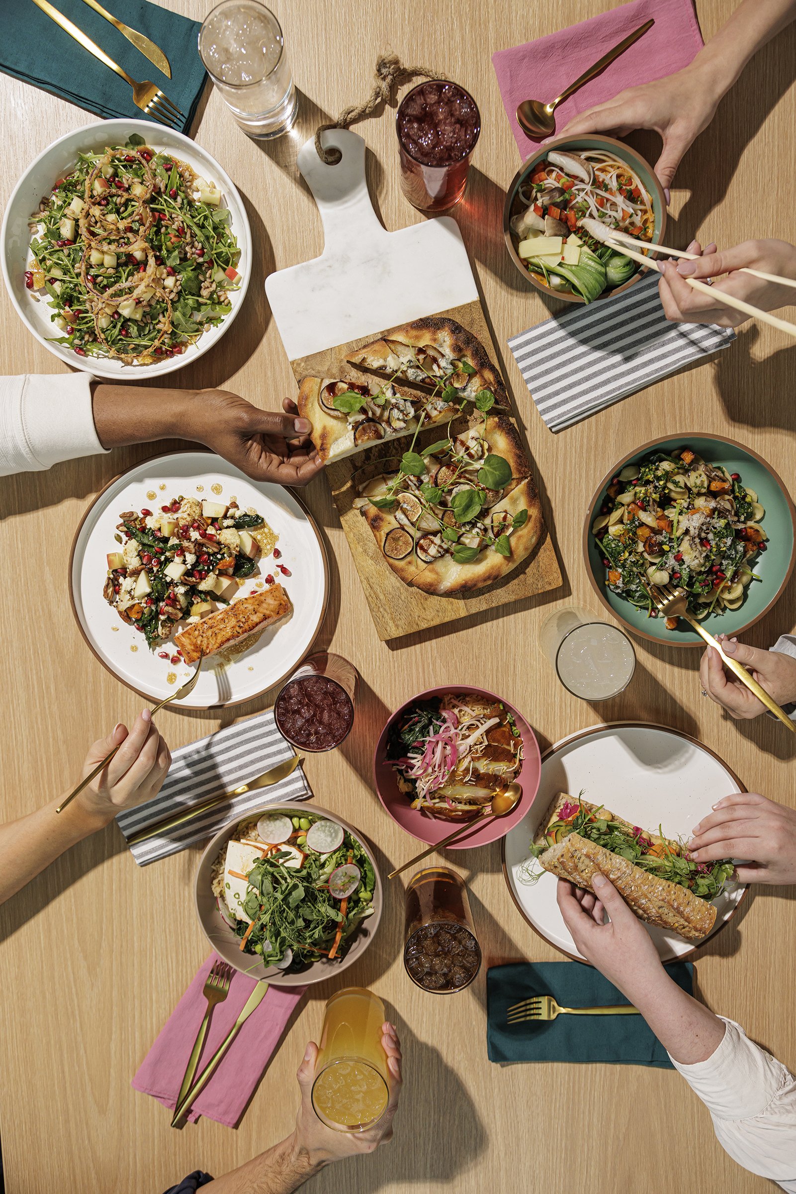 People sharing a meal with various dishes including pizza, salads, and bowls of Asian cuisine, along with drinks on a wooden table.