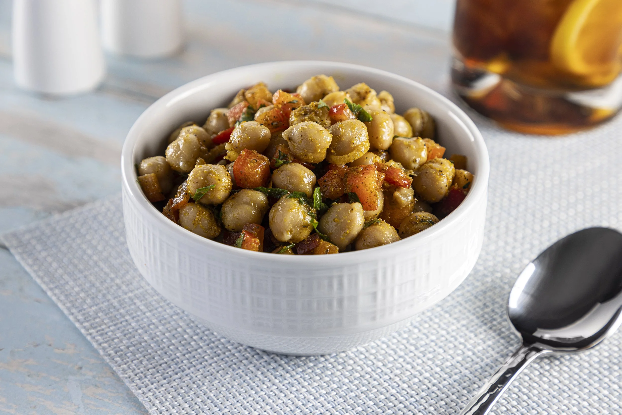 A white bowl filled with chickpea salad contains chickpeas, chopped tomatoes, and herbs. The bowl is on a white textured placemat on a light blue surface, with a spoon nearby and a jar with lemon slices and a dark liquid in the background.