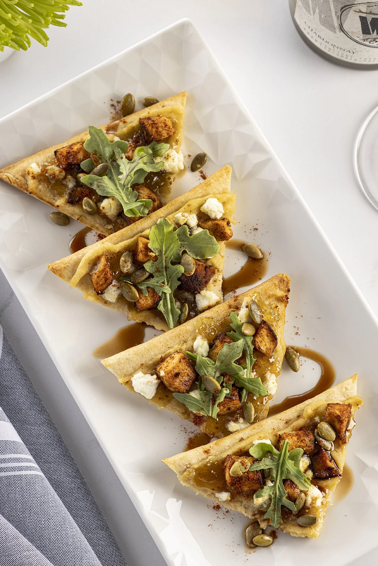 Top-down view of four slices of savory tart on a white rectangular plate, garnished with arugula, pumpkin seeds, crumbled cheese, and drizzled with a brown sauce.