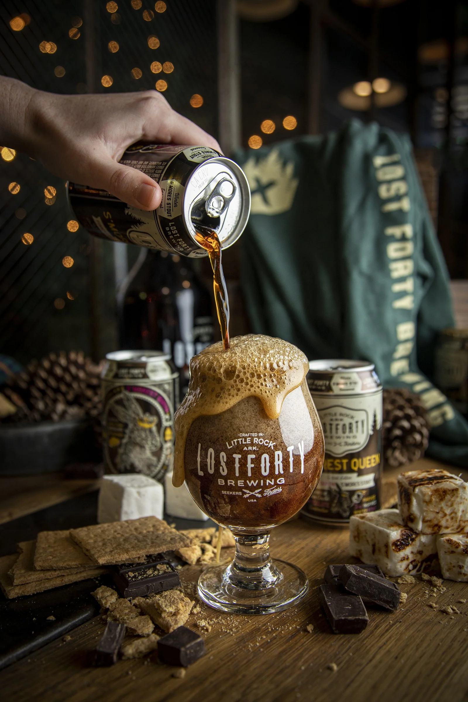 A hand pouring beer from a can into a branded beer glass, with assorted chocolates, cookies, and marshmallows on a wooden surface in the background.