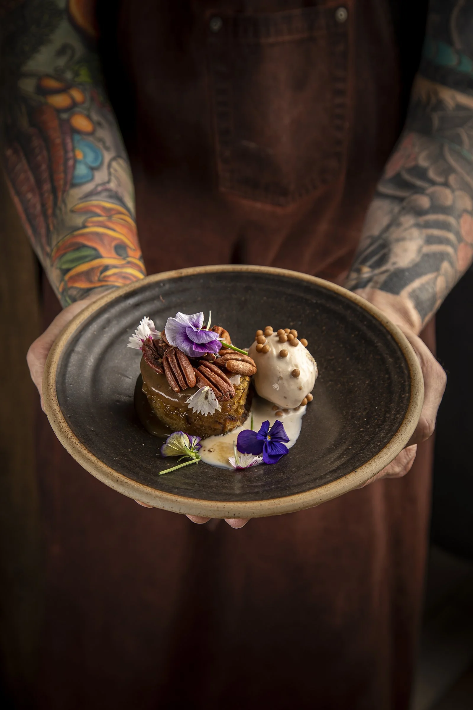 A person with colorful tattoos on their arms holding a dark ceramic plate with a dessert topped with pecans, edible flowers, and a scoop of ice cream.