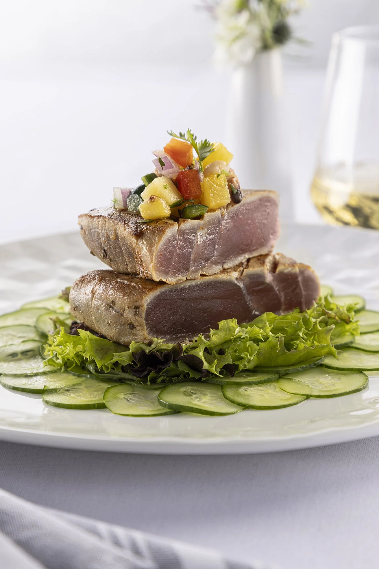Slices of seared tuna topped with diced pineapple, red bell pepper, purple onion, and a sprig of parsley, served on a bed of sliced cucumbers and assorted greens with a glass of white wine in the background.